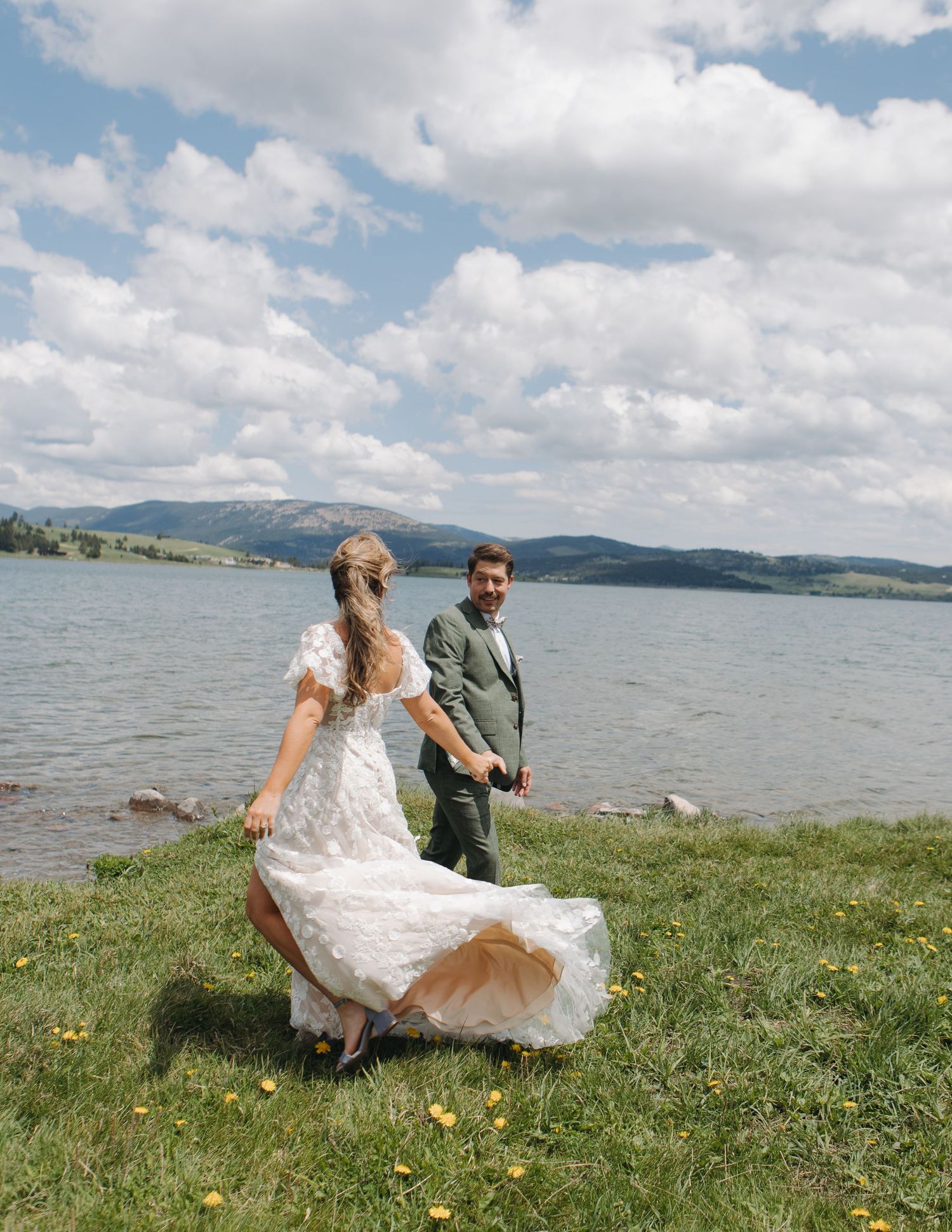 Wedding couple sitting on grassy hill overlooking lake and mountains on sunny day.