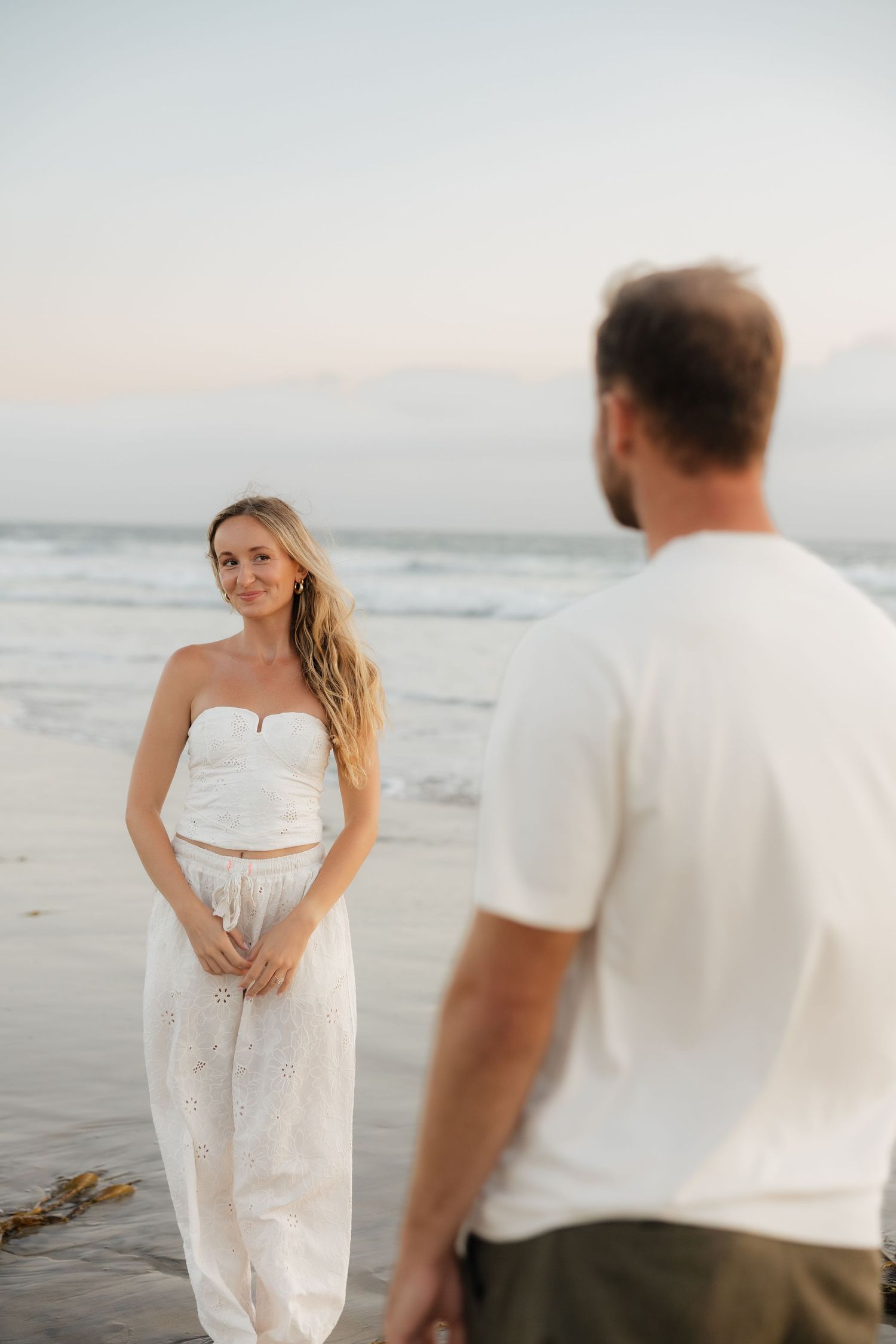 A woman in a white strapless top and skirt smiles at a man on a beach at sunset.