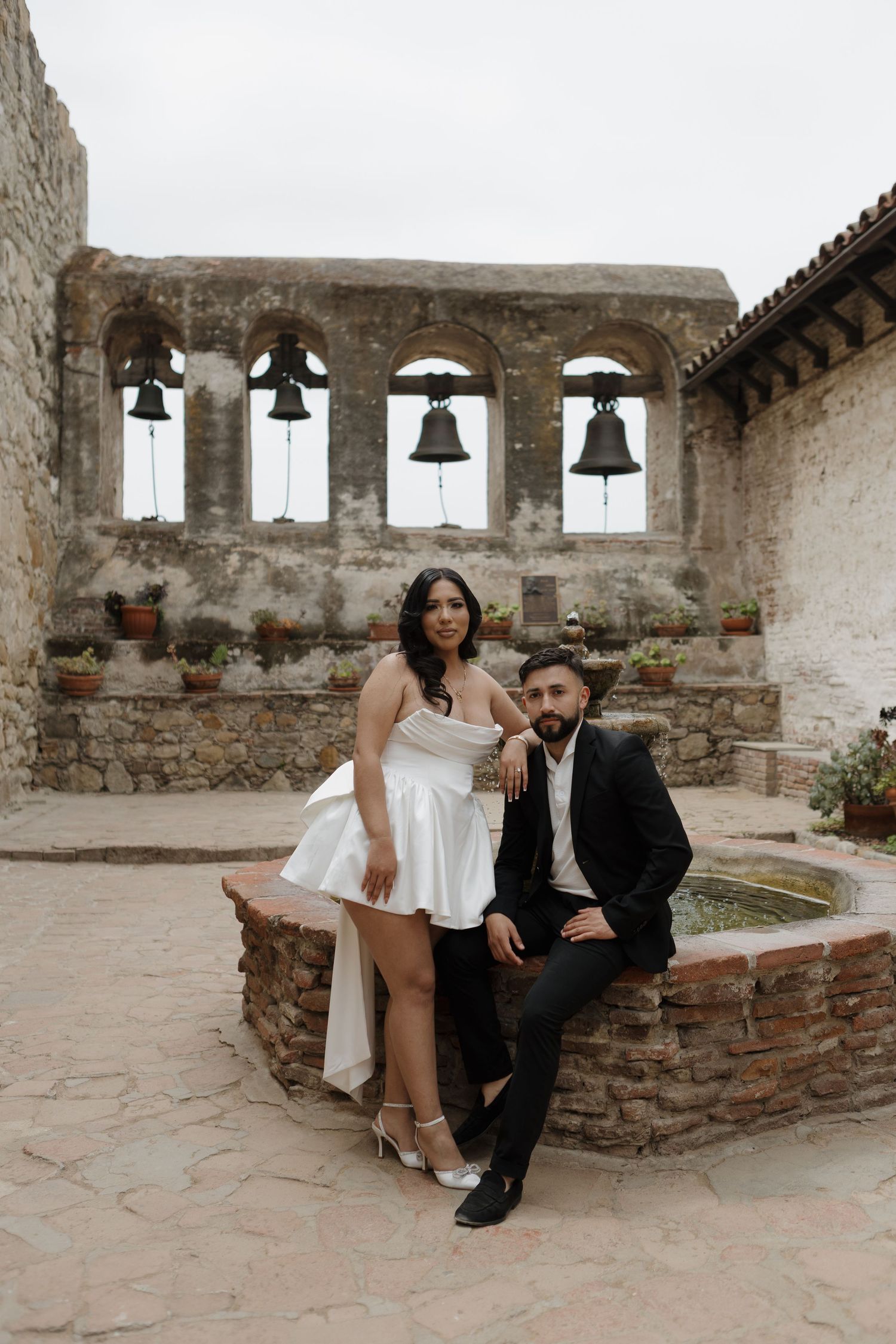 Couple poses in rustic mission courtyard with historic bells and stone walls.