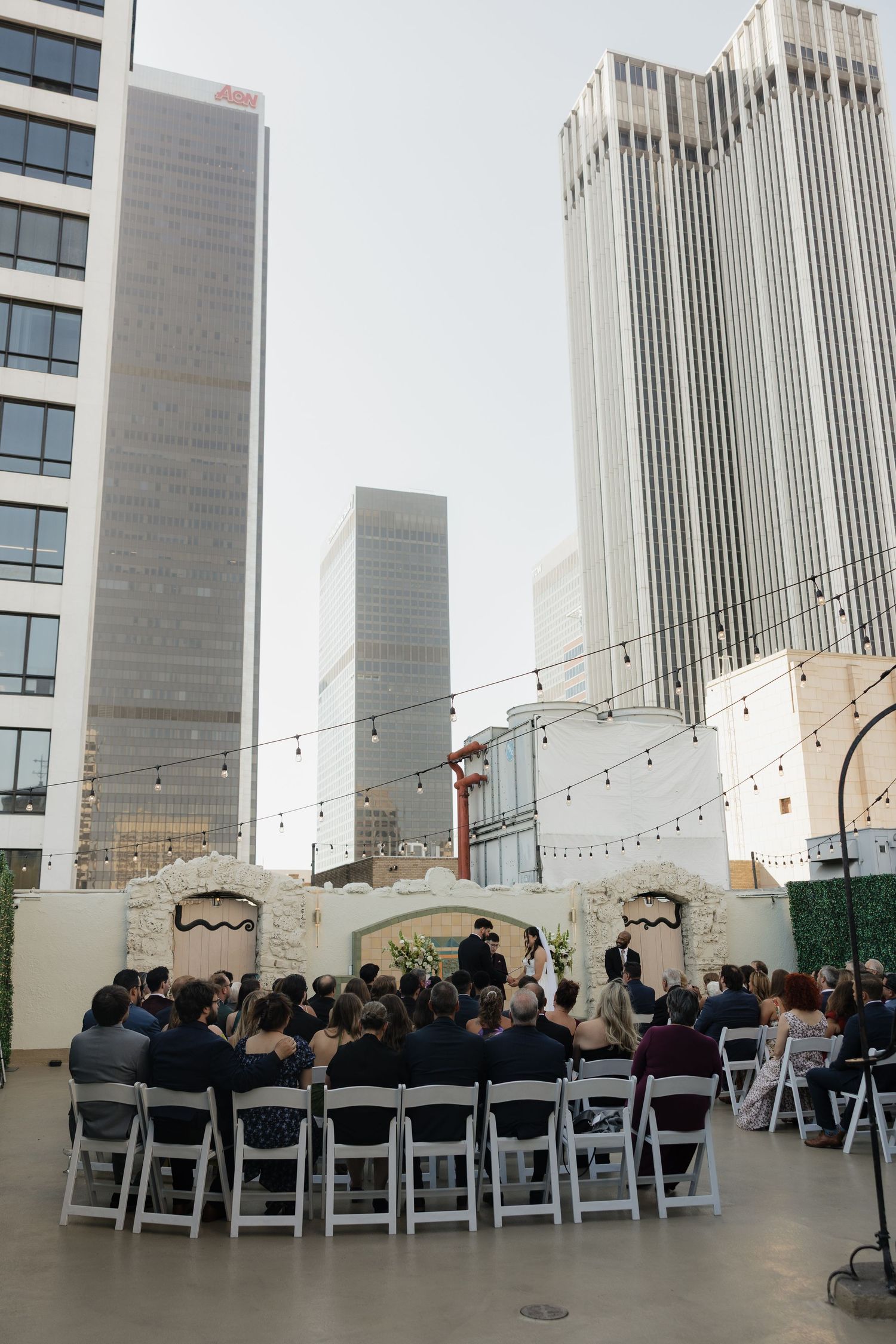 Outdoor rooftop wedding ceremony with white chairs and city skyscrapers backdrop under string lights.