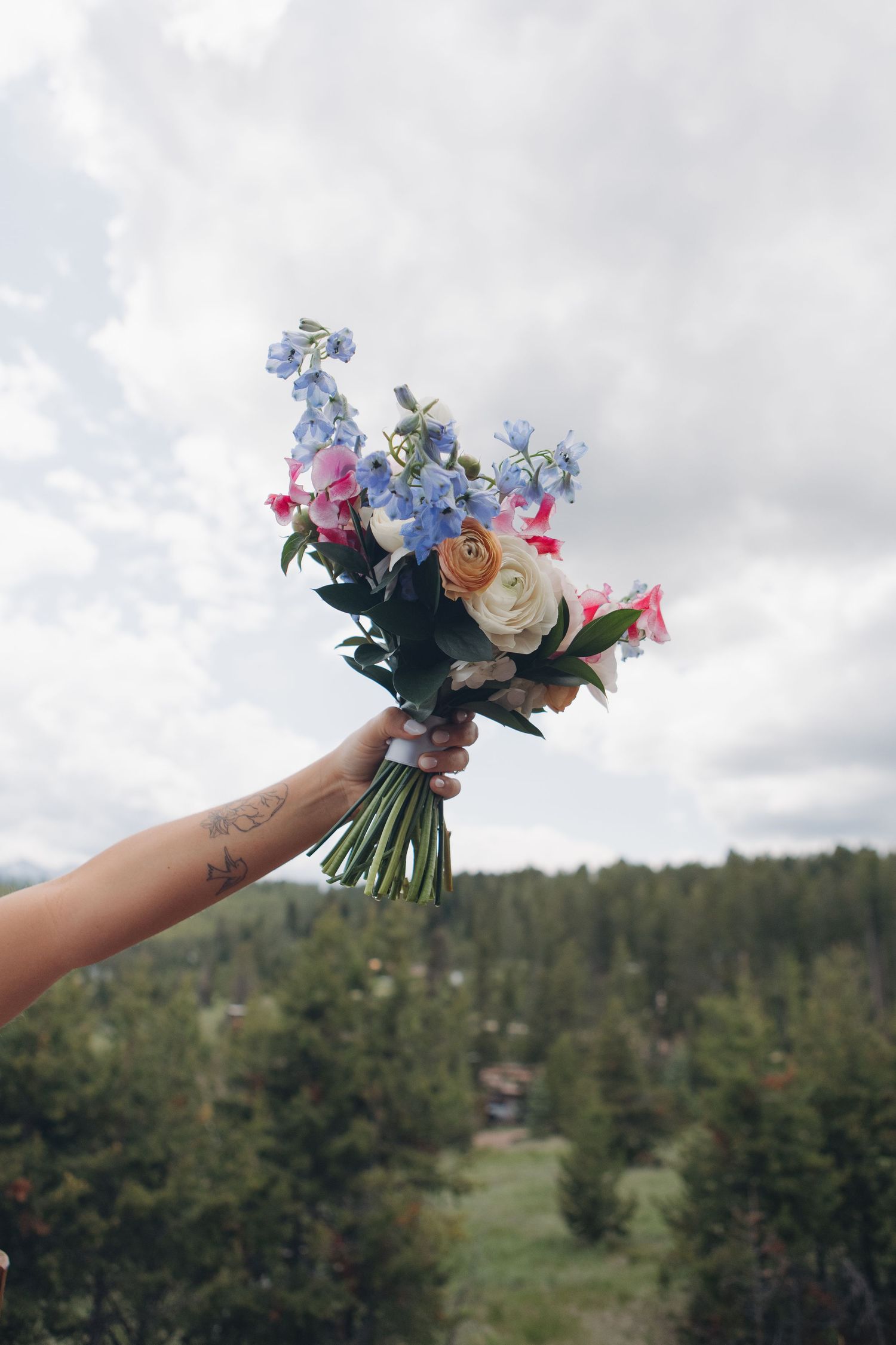 Hand holding a vibrant bridal bouquet with blue and pink flowers against a mountain landscape.