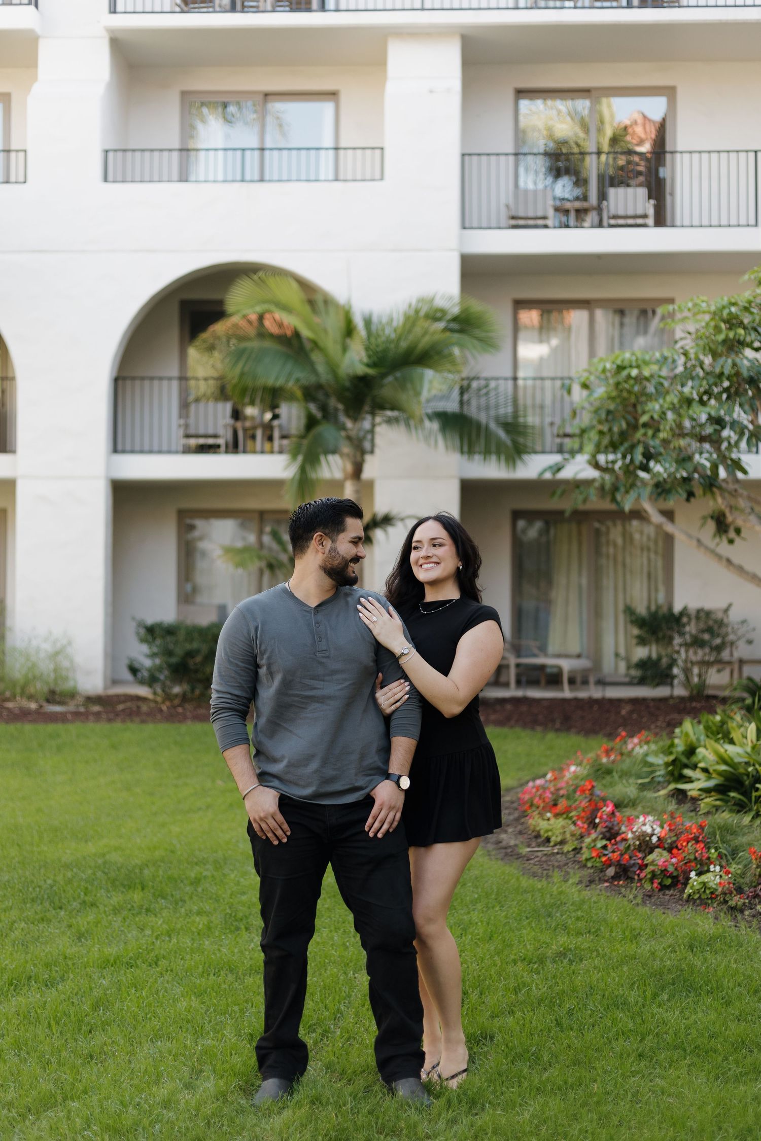 Couple in black attire playfully posing on grass in front of apartment complex.