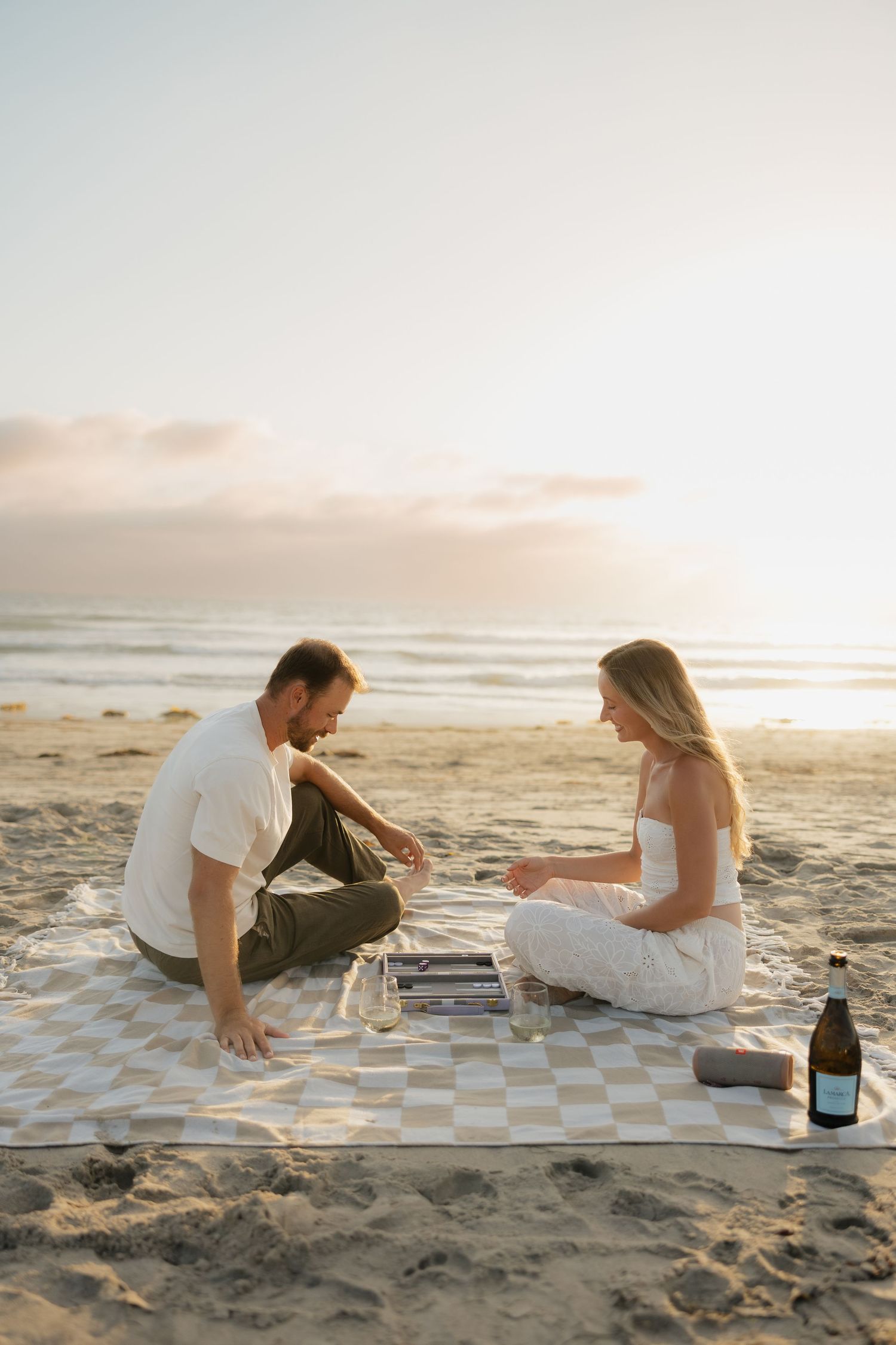Couple playing backgammon on a checkered blanket at the beach during golden hour sunset.