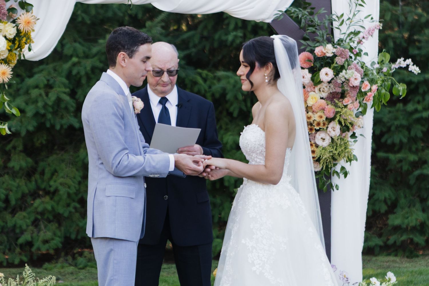 A wedding ceremony takes place under a white floral arch in a garden setting.