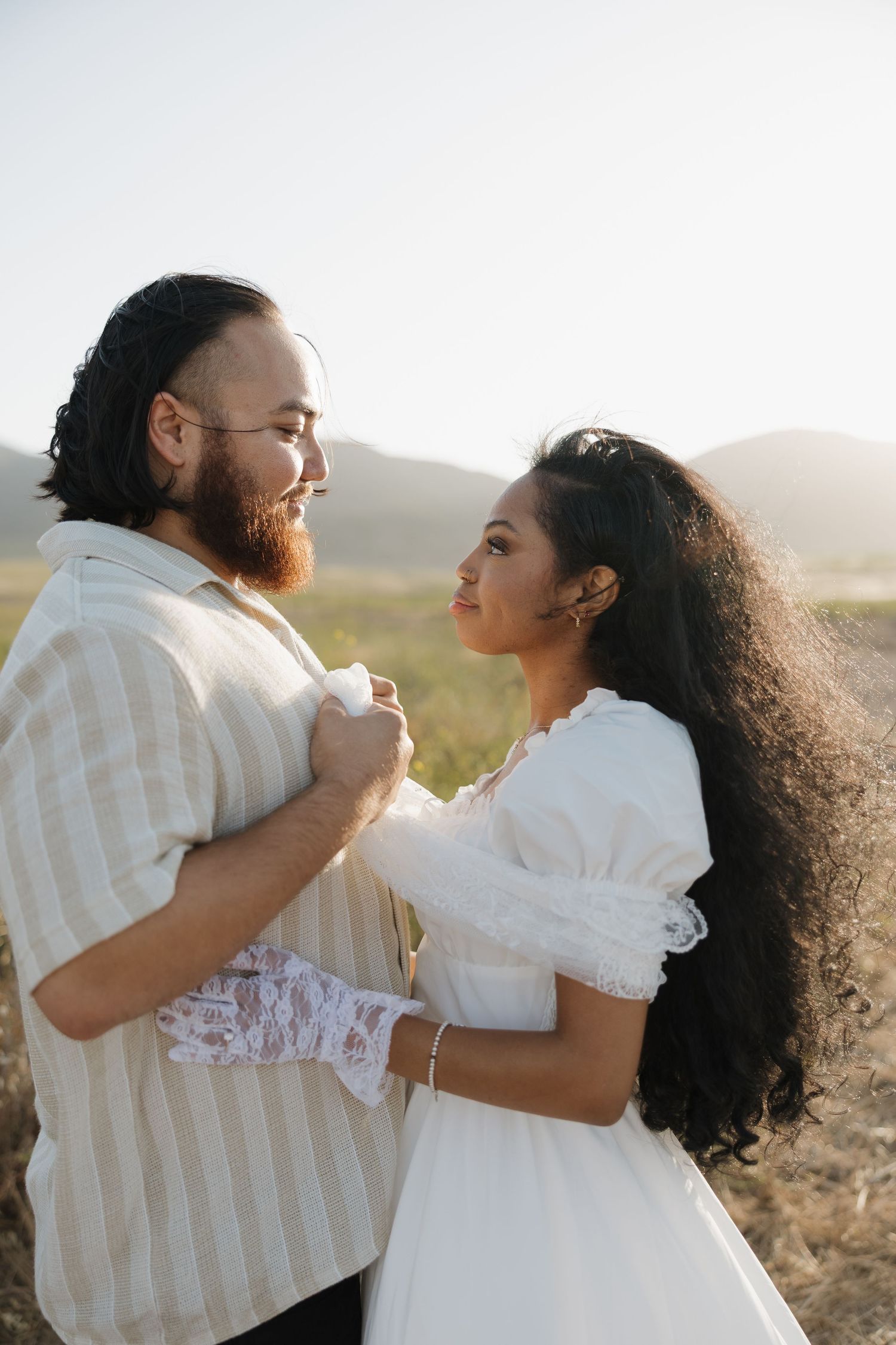 A couple in boho attire embraces in a sunlit field with mountains in the background.