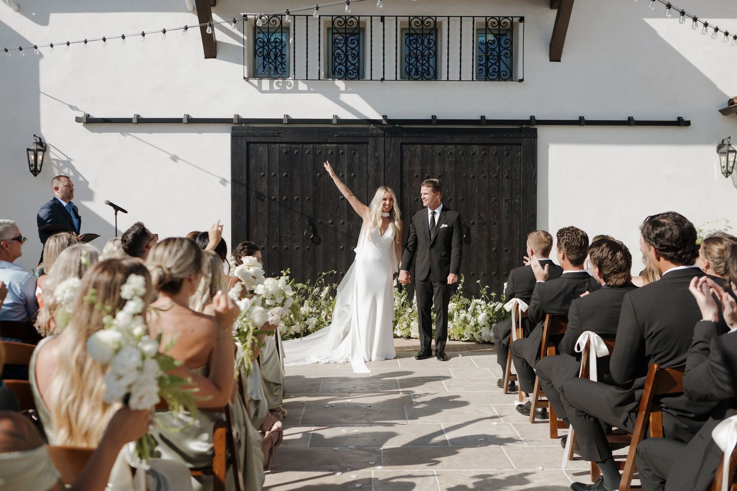 Wedding party celebrates in outdoor courtyard with string lights and white architecture.