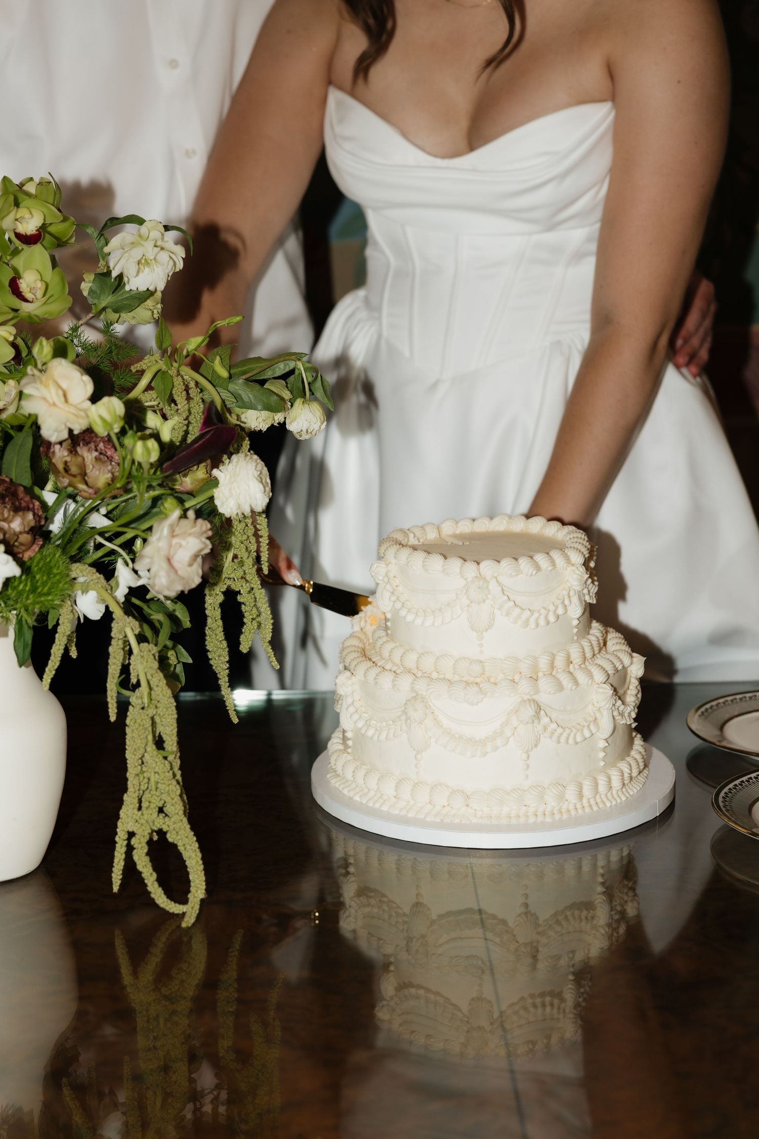 Bride in white strapless gown cutting a two-tier white wedding cake beside a floral arrangement.
