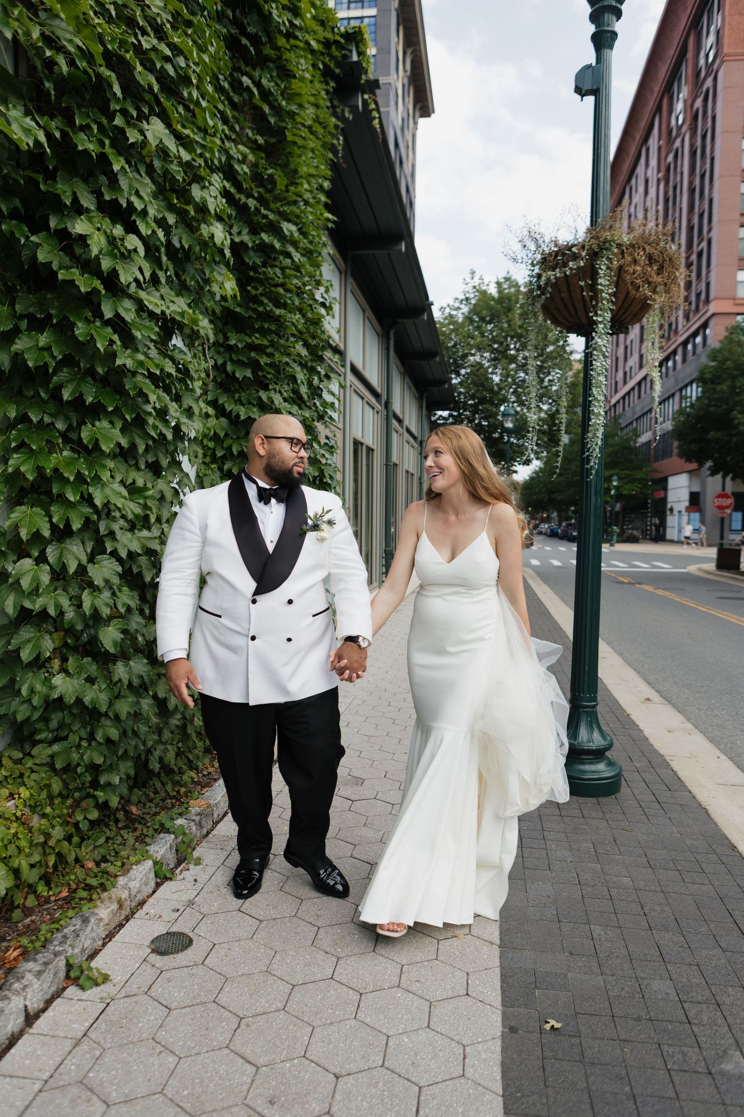 Bride and groom walk hand-in-hand on a city sidewalk, groom in white tuxedo, bride in sleek white gown.