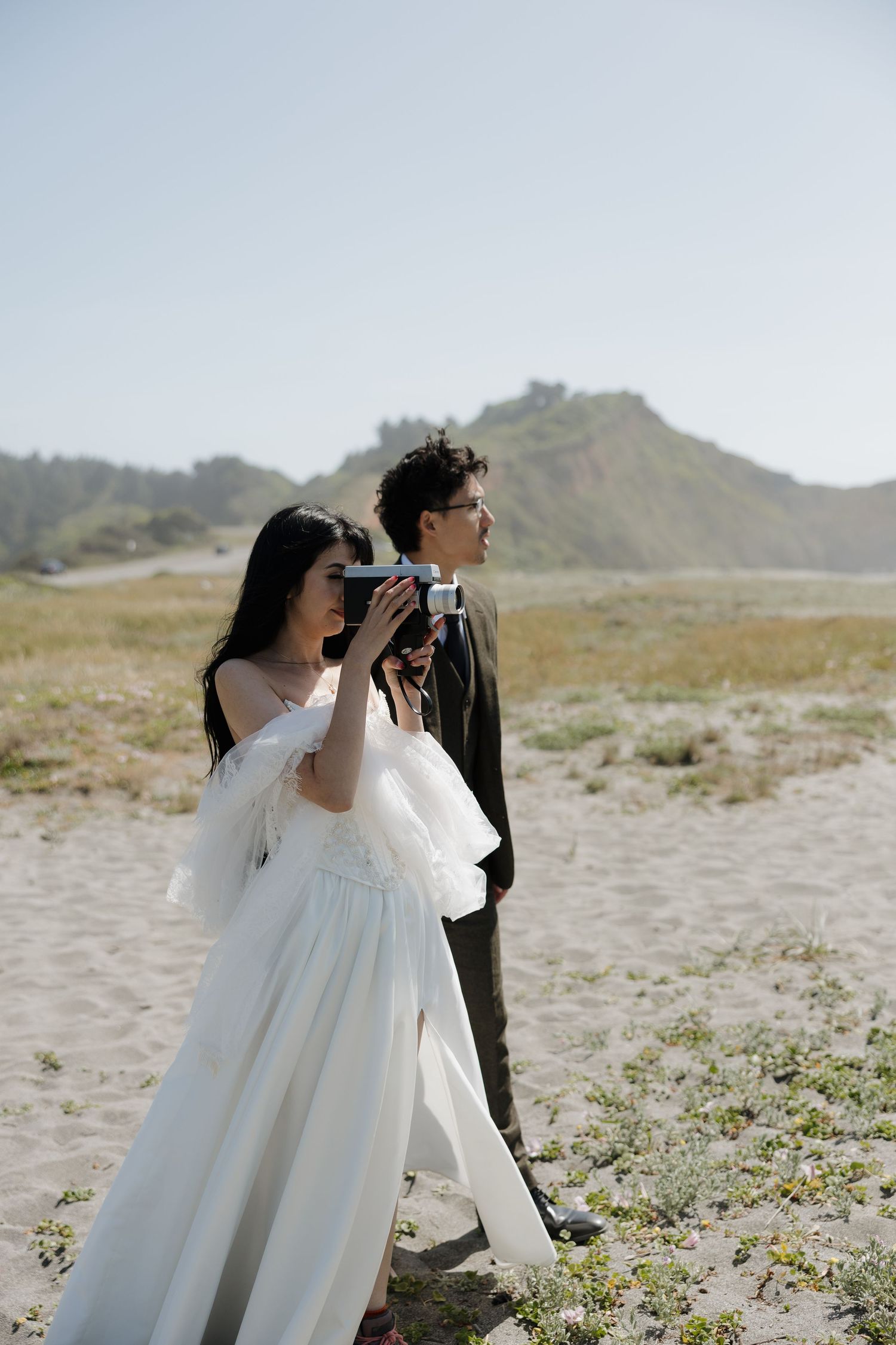 Couple in wedding attire on sandy beach, bride in white gown holding vintage camera, green hills in background.