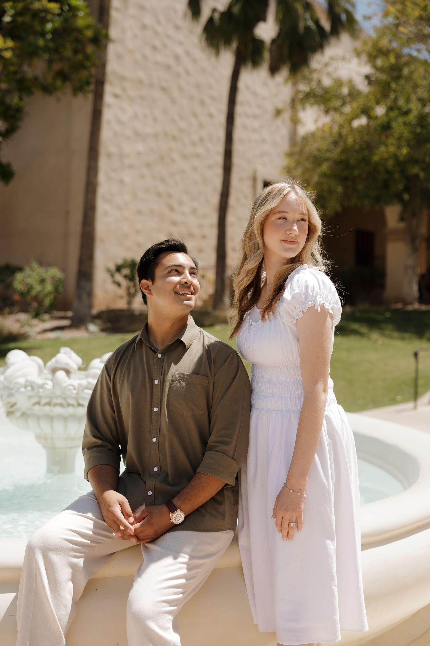 A couple poses by a fountain outdoors, man in olive shirt seated, woman in white dress standing, palm trees behind.