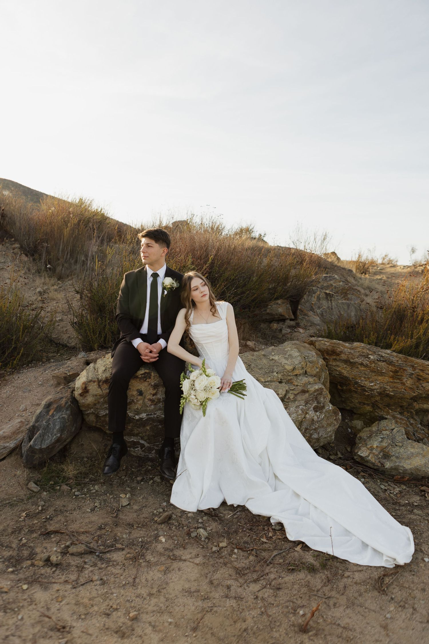 Couple in wedding attire sits on rocky terrain at sunset.