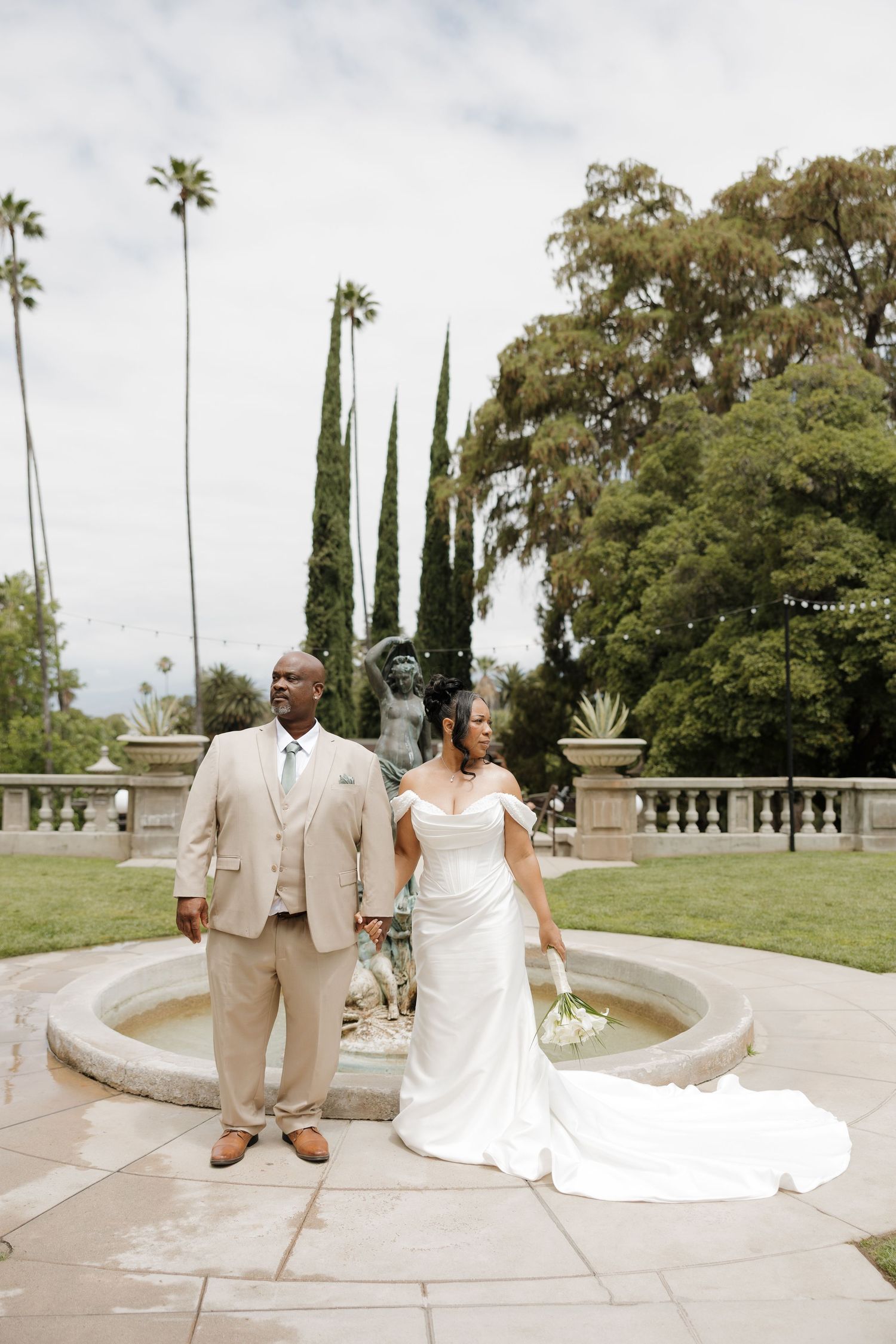 Bride and groom pose by a fountain in a garden with palm trees, she in a white gown, he in a tan suit.