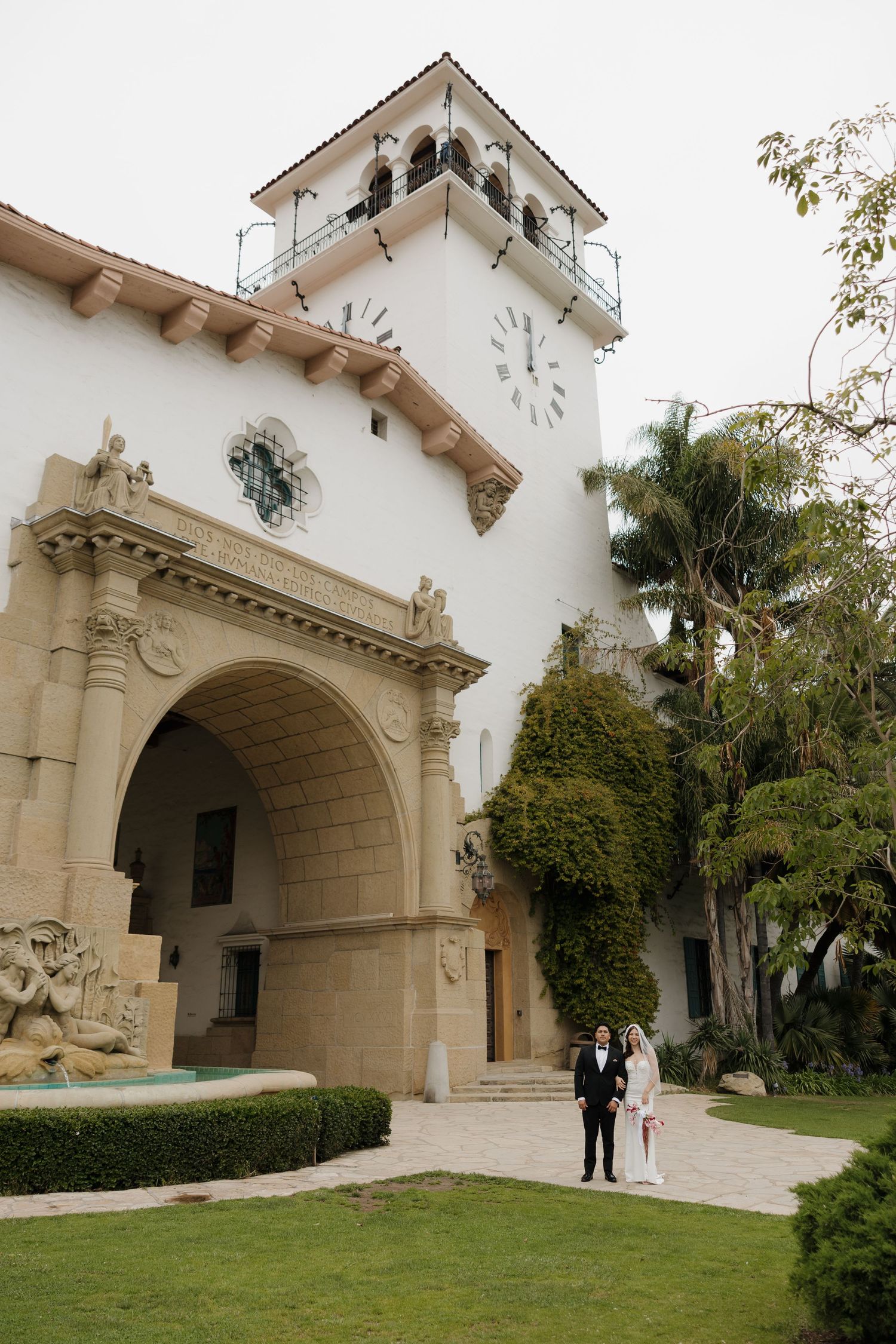 Bride and groom stand before Santa Barbara Courthouse's ornate Spanish Colonial archway and clock tower surrounded by lush gardens.