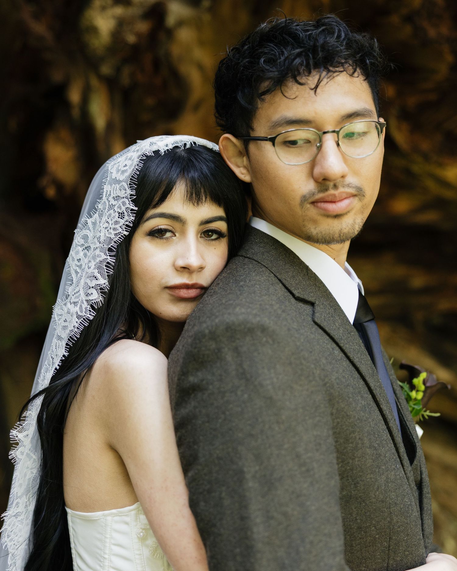 Bride in white gown and lace veil poses back-to-back with groom in brown suit during outdoor wedding portrait.