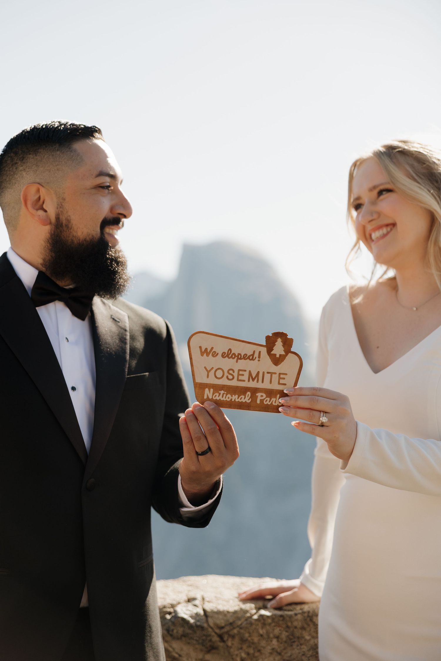 Couple in wedding attire smiling and holding a wooden Yosemite National Park elopement sign outdoors.