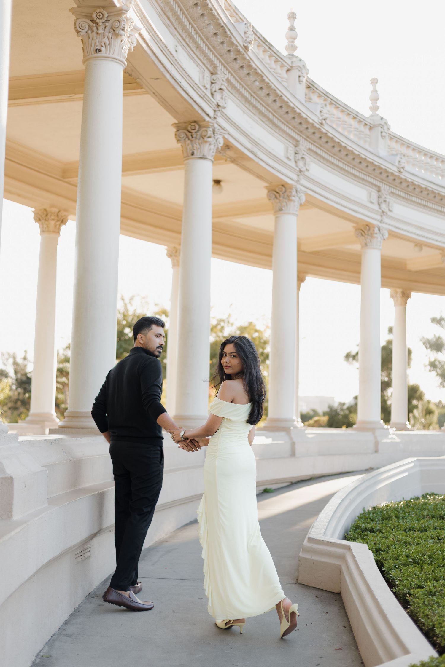 A couple in wedding attire holding hands at an elegant white columned pavilion.
