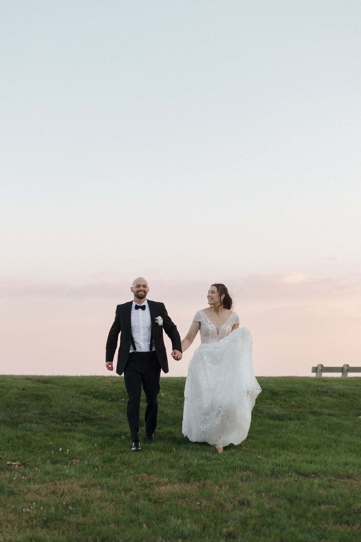 A couple runs playfully across a grassy hillside at sunset in formal wedding attire.