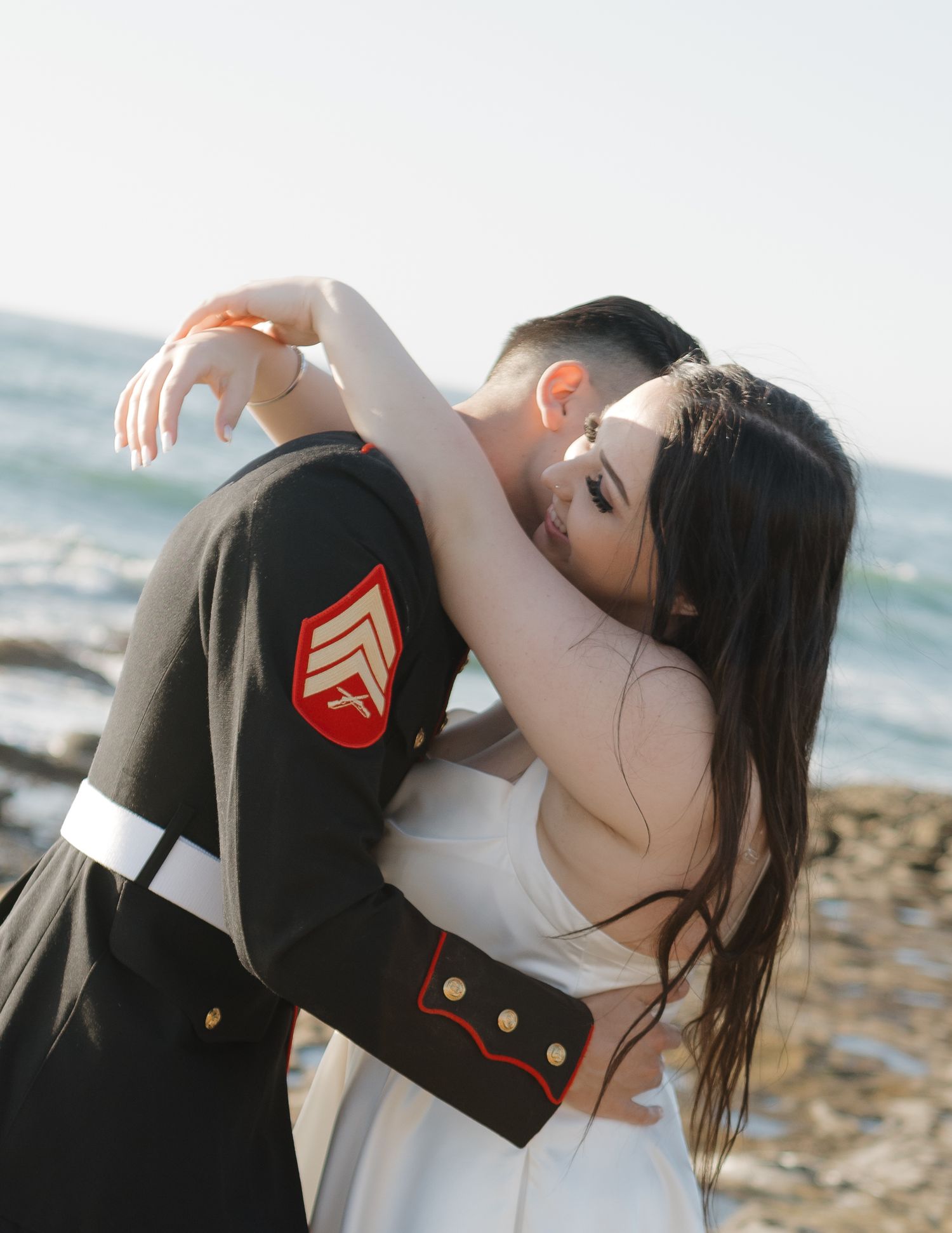 A romantic embrace captured on a sunny beach with military dress uniform details.