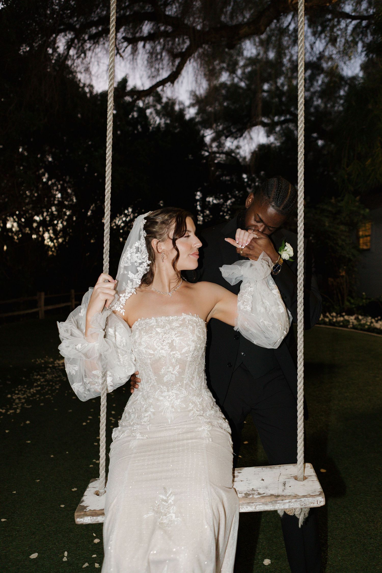 Bride in lace gown on rustic swing as groom in black suit kisses her head outdoors at dusk.