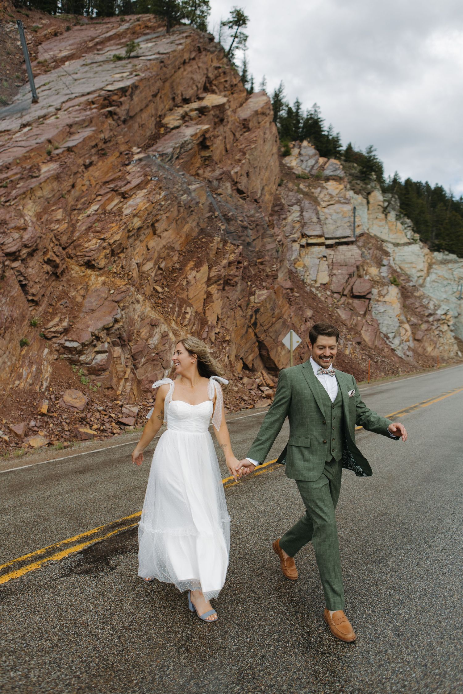 A couple playfully runs and dances on a mountain road with rocky cliffs in the background.