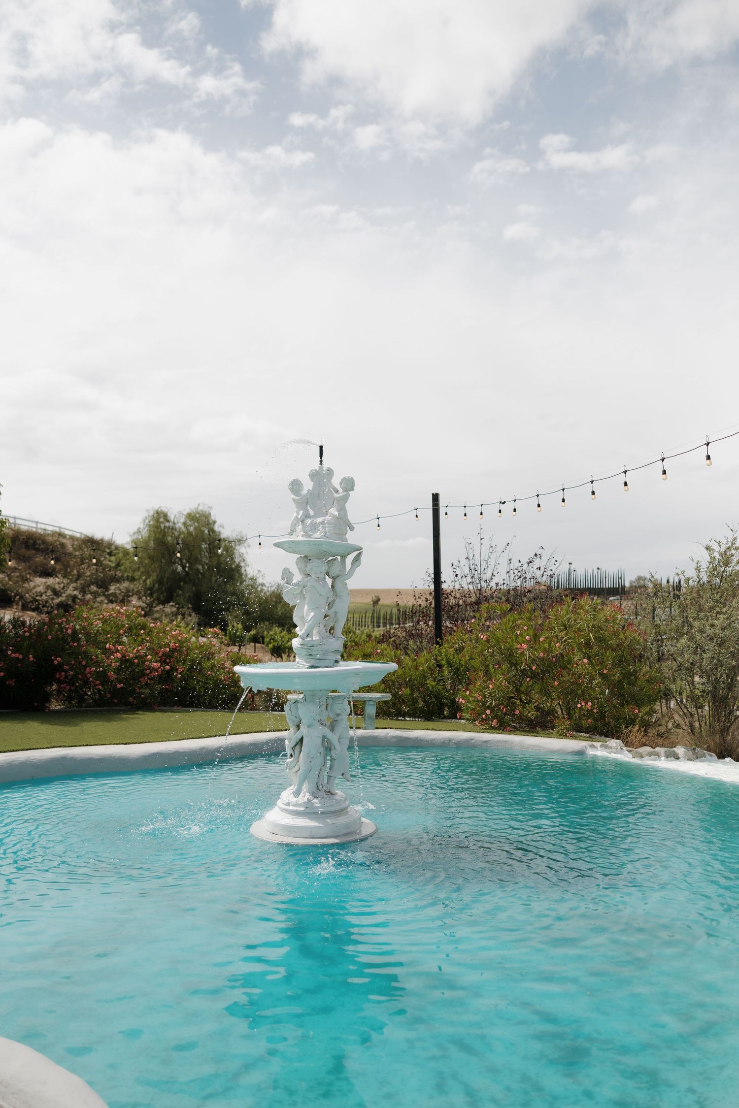 White ornate tiered fountain in a blue pool surrounded by lush gardens and string lights under a cloudy sky.