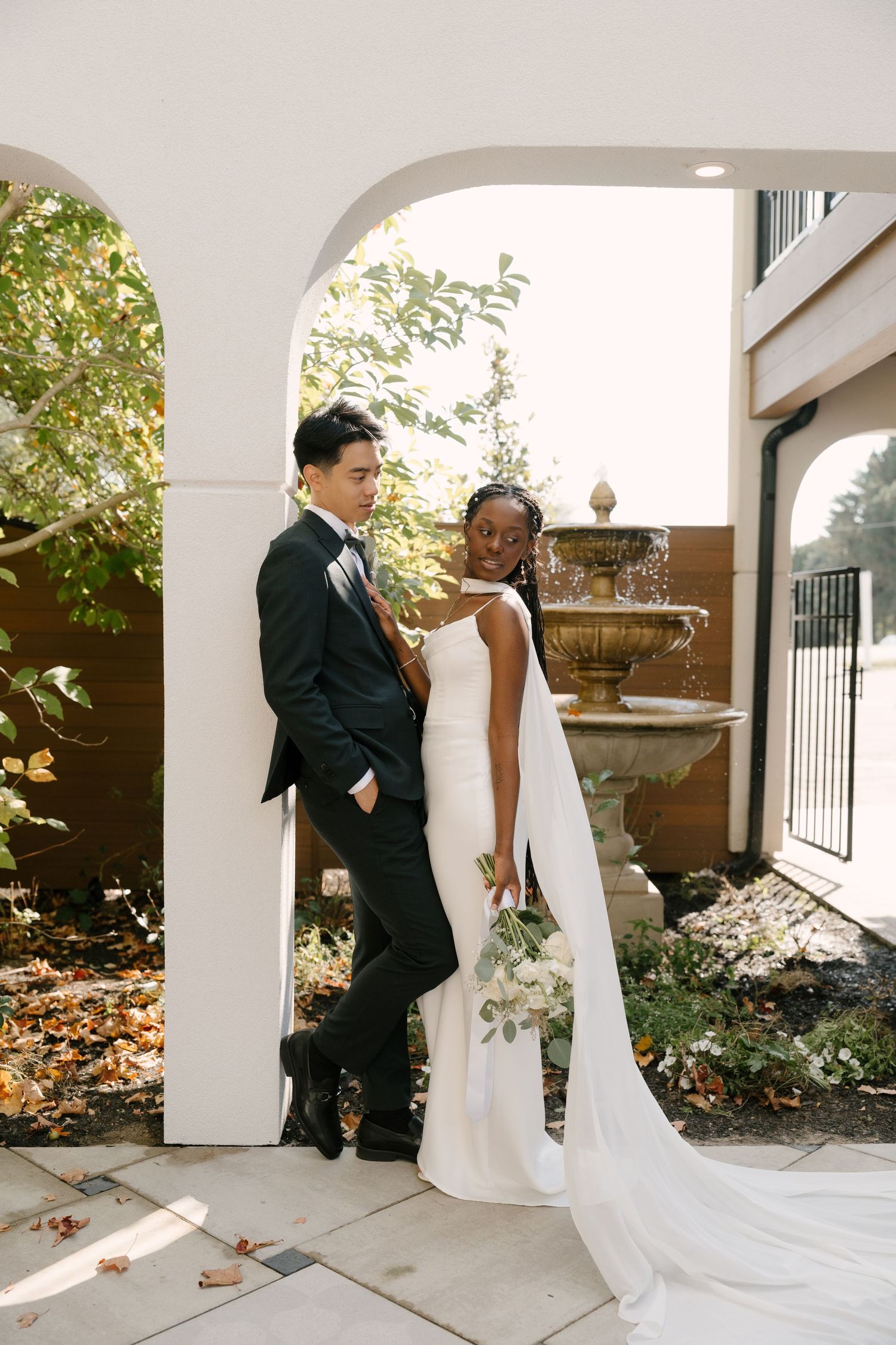 Interracial wedding couple posing under white arch with fountain backdrop in autumn garden setting at Wren House wedding venue.