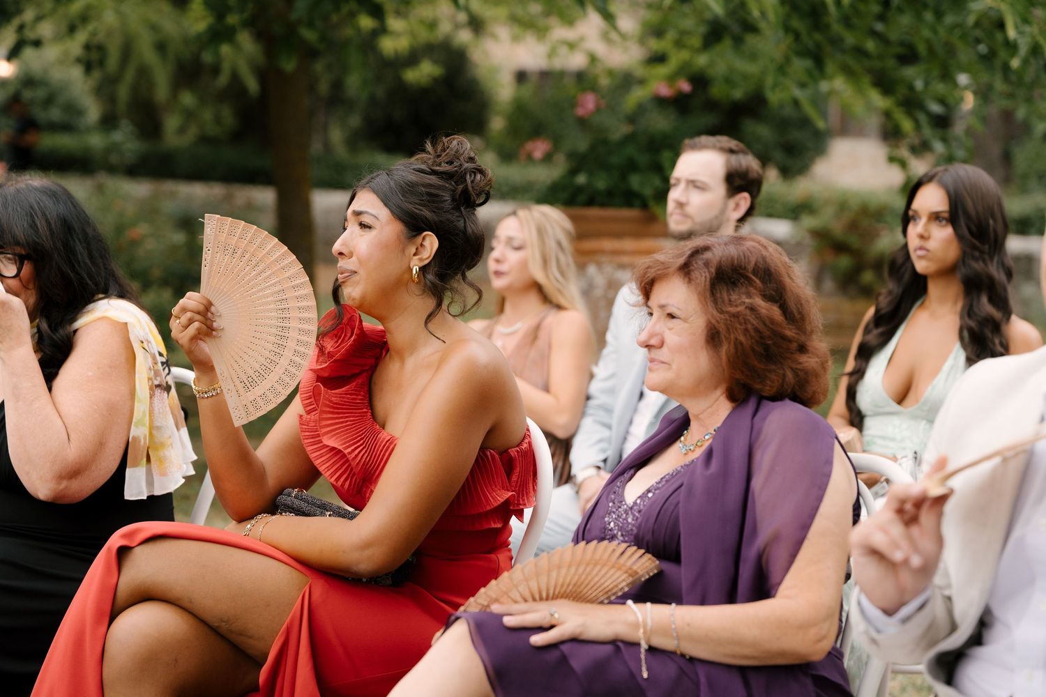 Wedding guests seated outdoors, fanning themselves in the heat, wearing elegant attire at a garden ceremony.