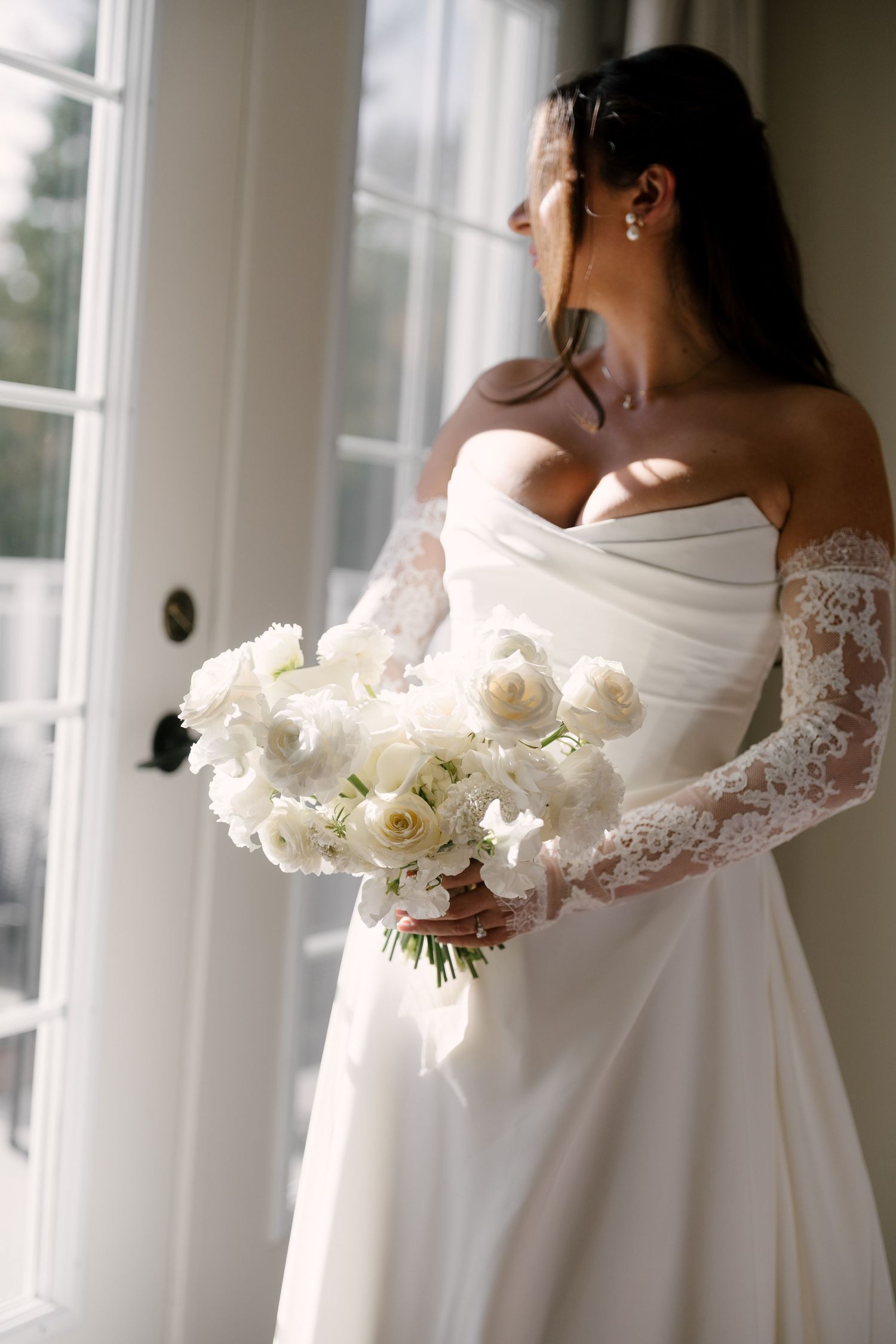 Bride in strapless gown with lace sleeves holds white floral bouquet near sunlit French doors.