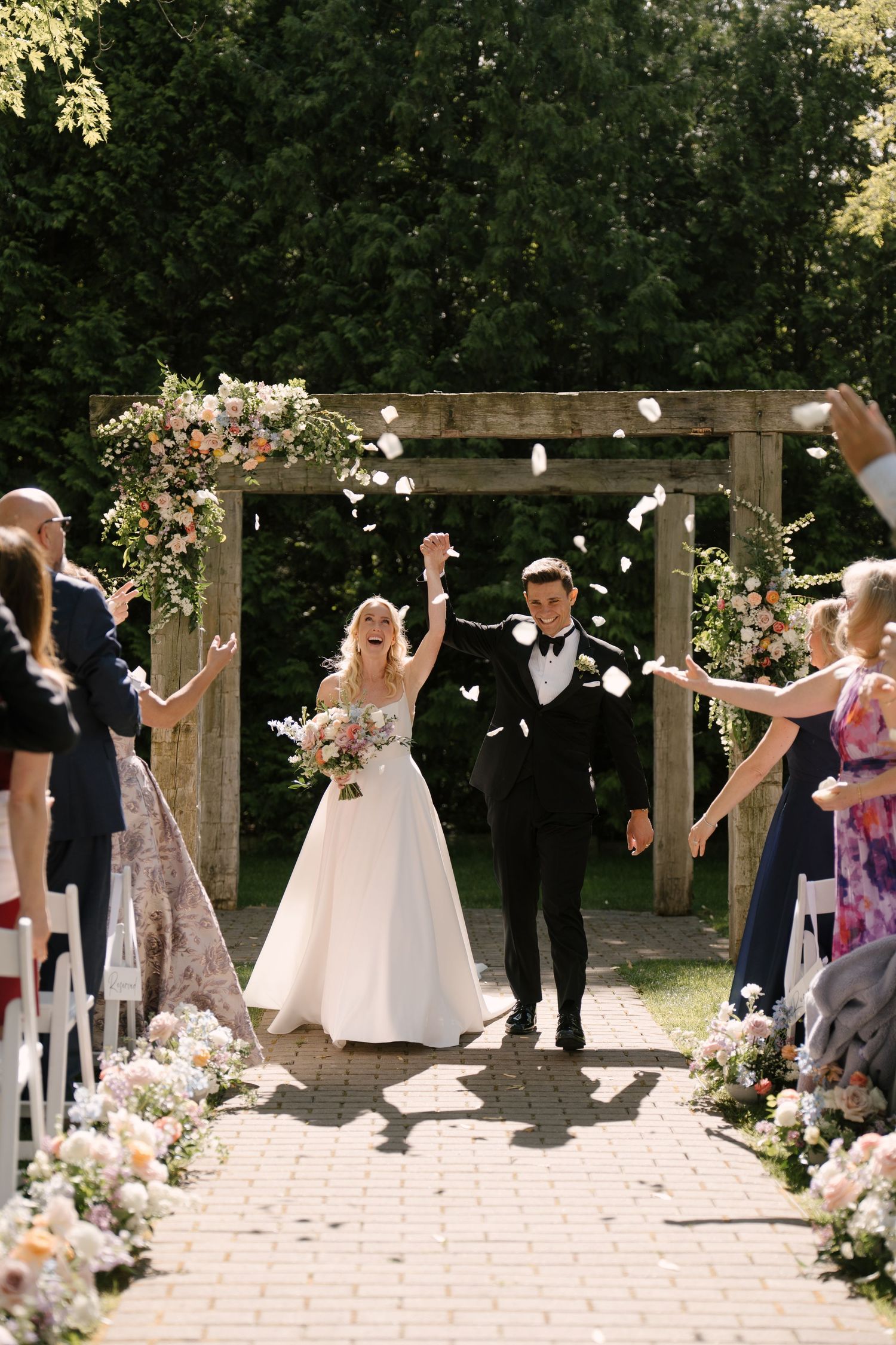 Bride and groom walk down outdoor aisle as guests toss flower petals under wooden arch with floral decor at Roseville Estates wedding venue.