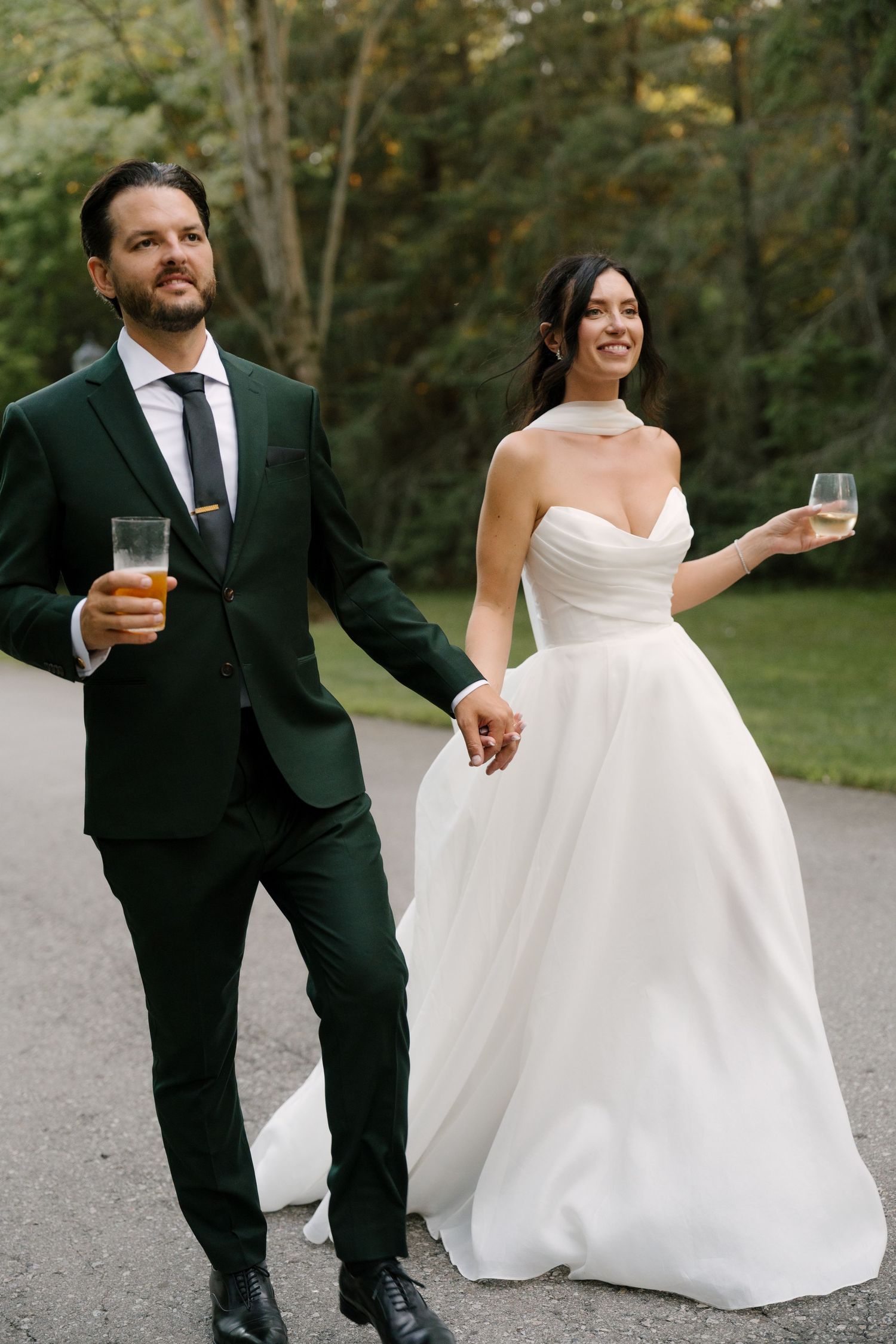 Bride and groom holding champagne glasses while walking hand in hand on a path surrounded by trees at Roseville Estates.