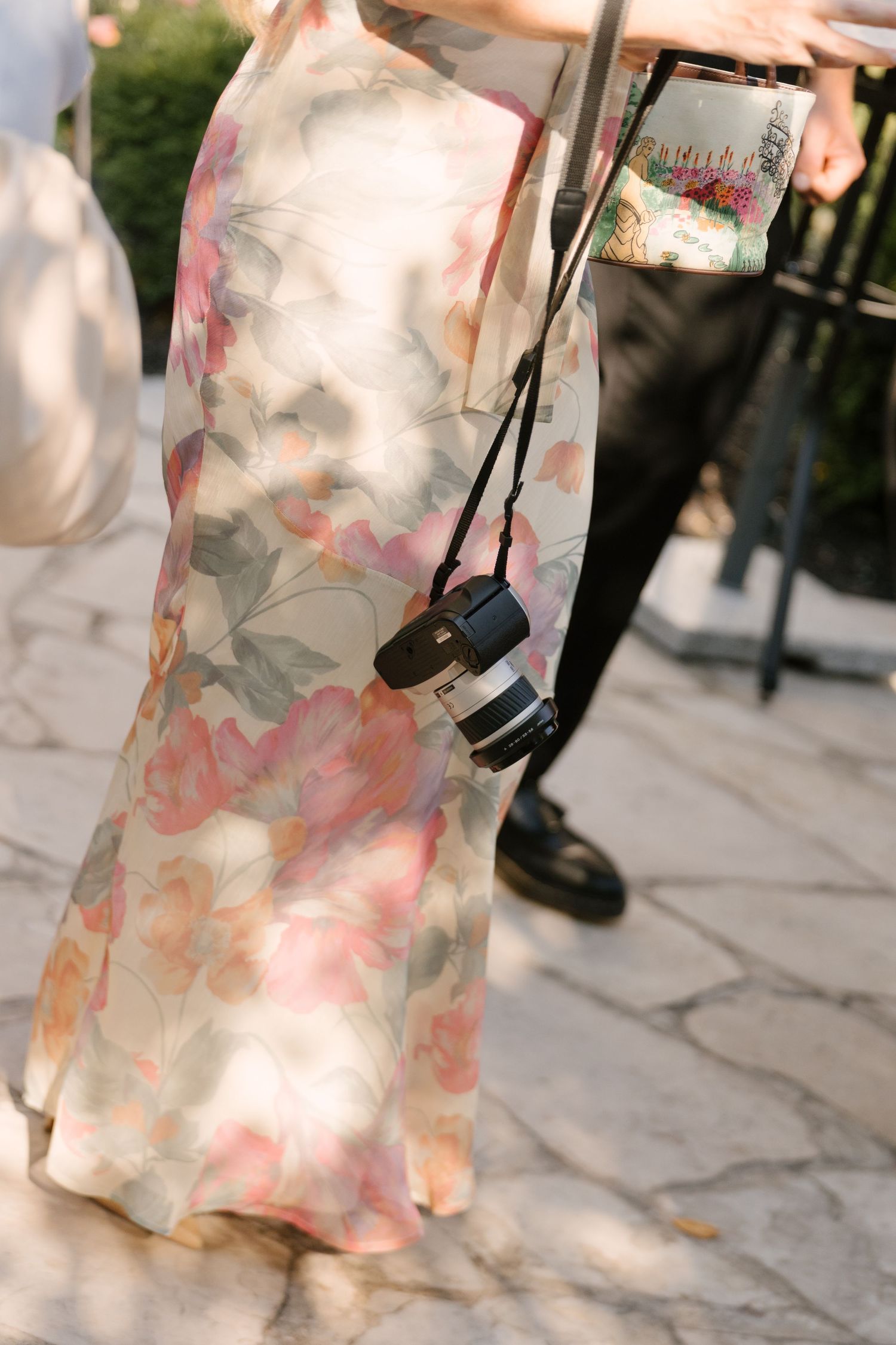 Wedding guest in floral maxi dress carrying a camera on strap at outdoor cocktail hour.