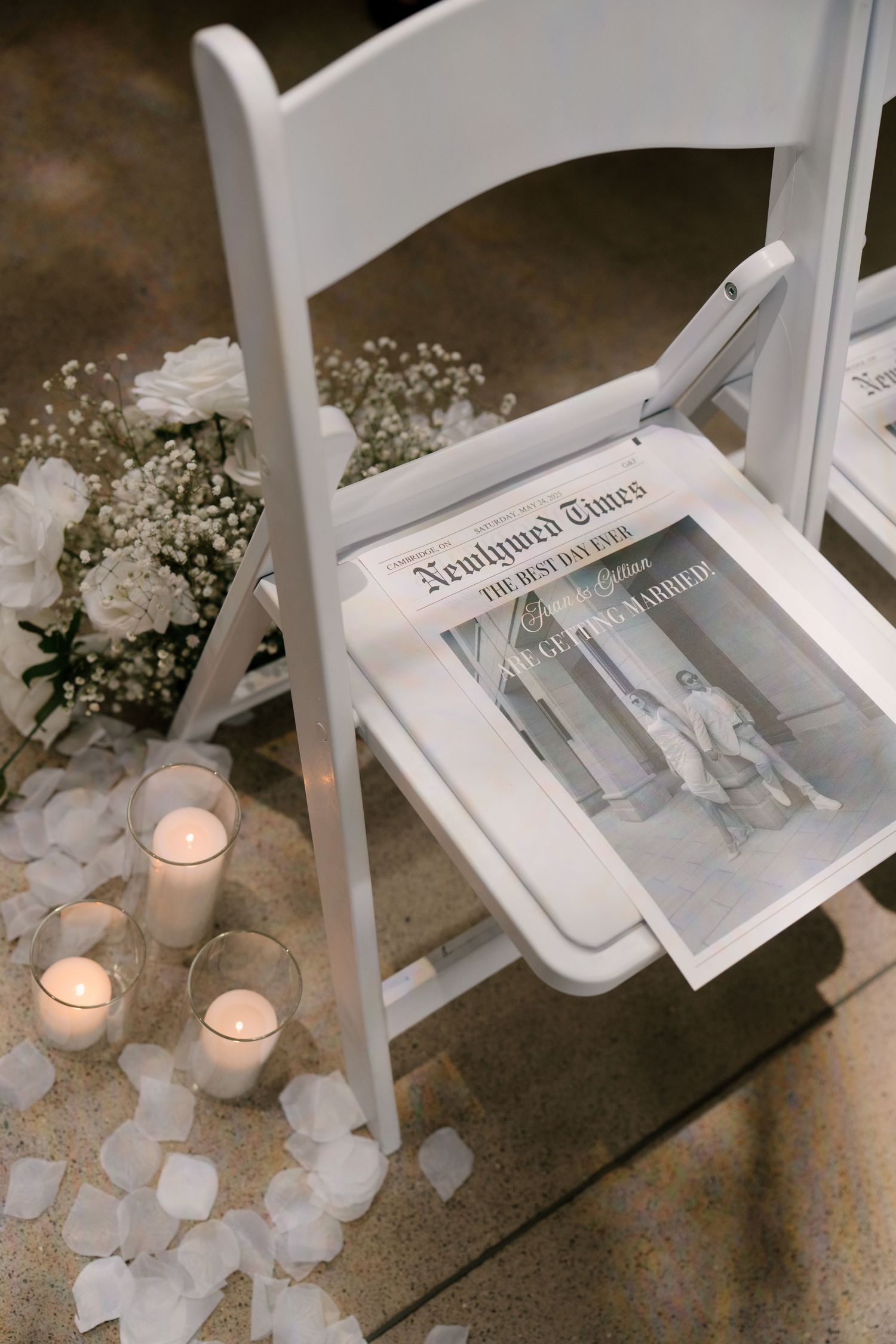 White folding chair with Newlywed Times program surrounded by candles and white flowers on a petal-strewn floor.
