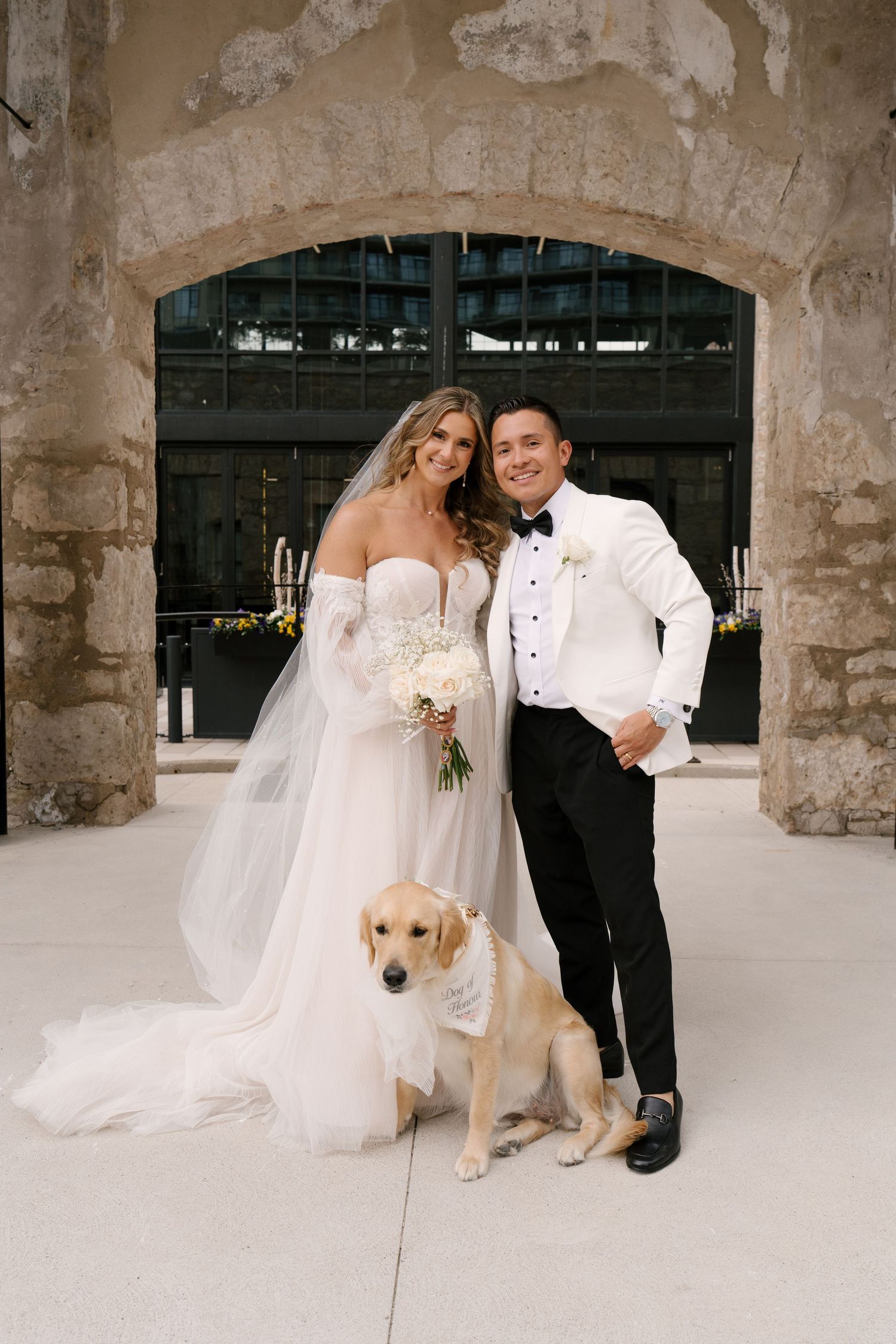 Bride and groom pose with golden retriever dog wearing white collar at stone arch wedding venue.