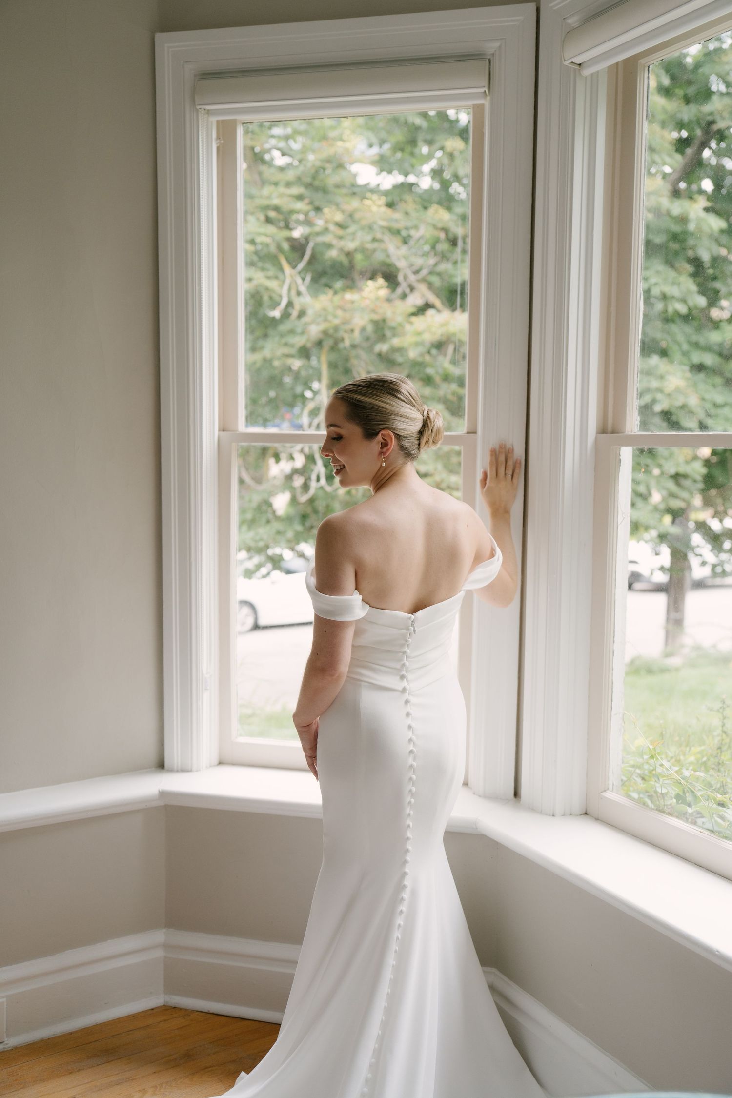 Bride in off-shoulder mermaid wedding gown with train gazing out a bay window.