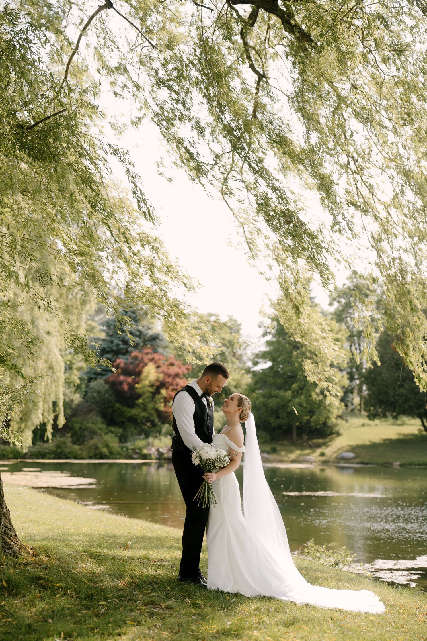 Bride and groom share a tender moment under a weeping willow tree beside a serene pond.