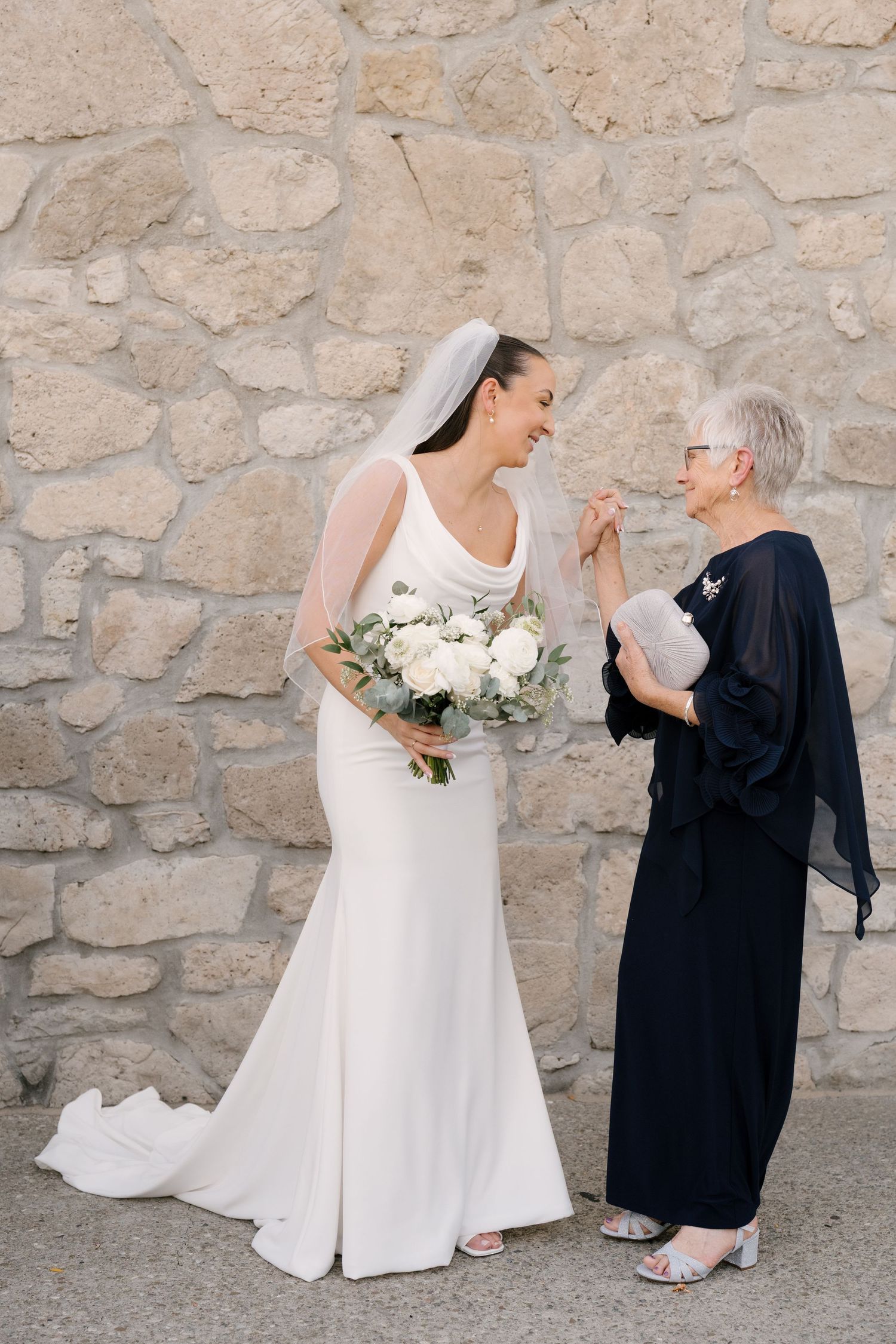 Bride in white fitted gown with veil holding white bouquet, laughing with elderly woman in navy dress outdoors at Cambridge Mill.
