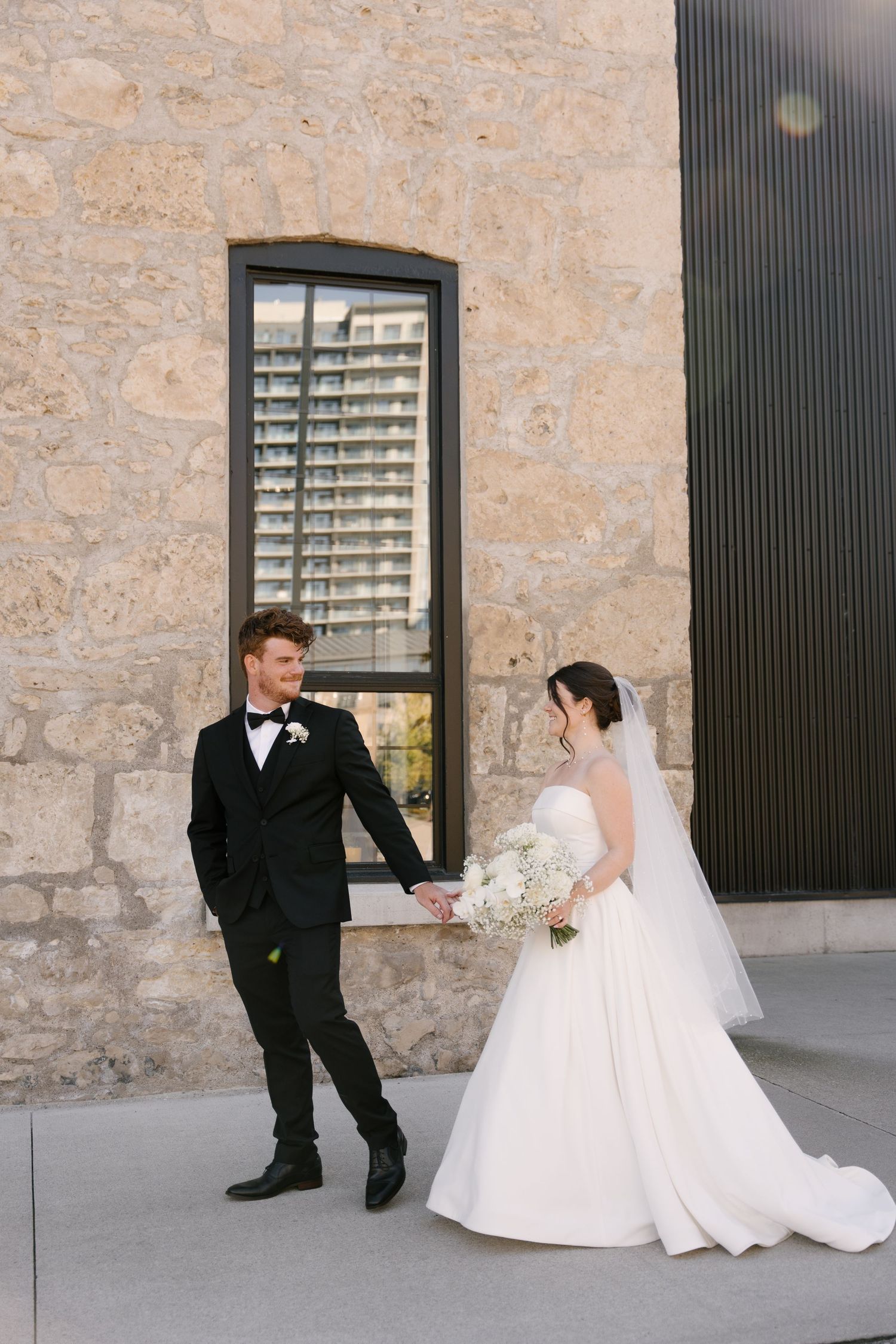 Bride in strapless ball gown and groom in black tuxedo hold hands by stone Tapestry Hall building.