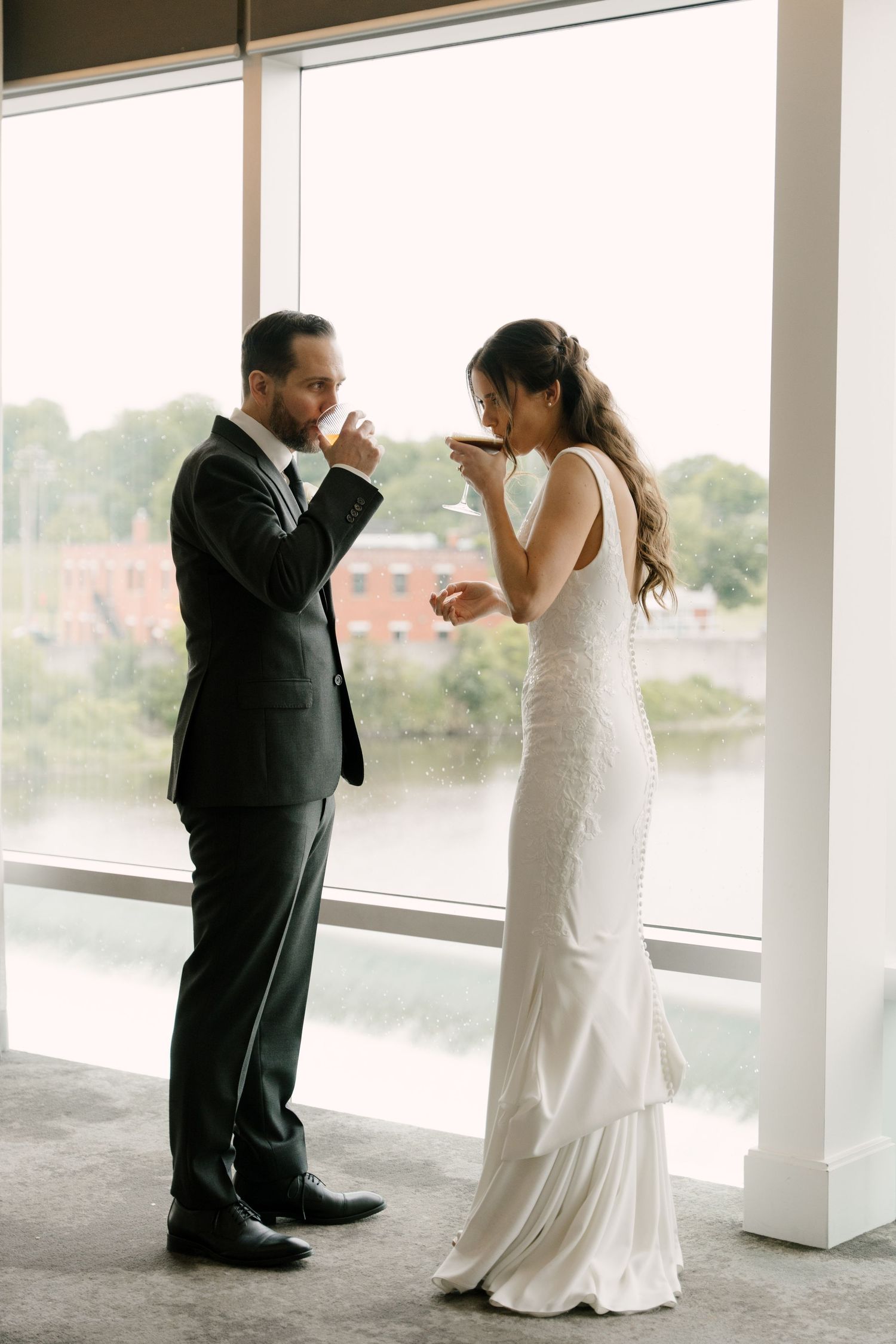 Bride and groom share a ceremonial drink by large windows overlooking a scenic river view at Cambridge Mill.
