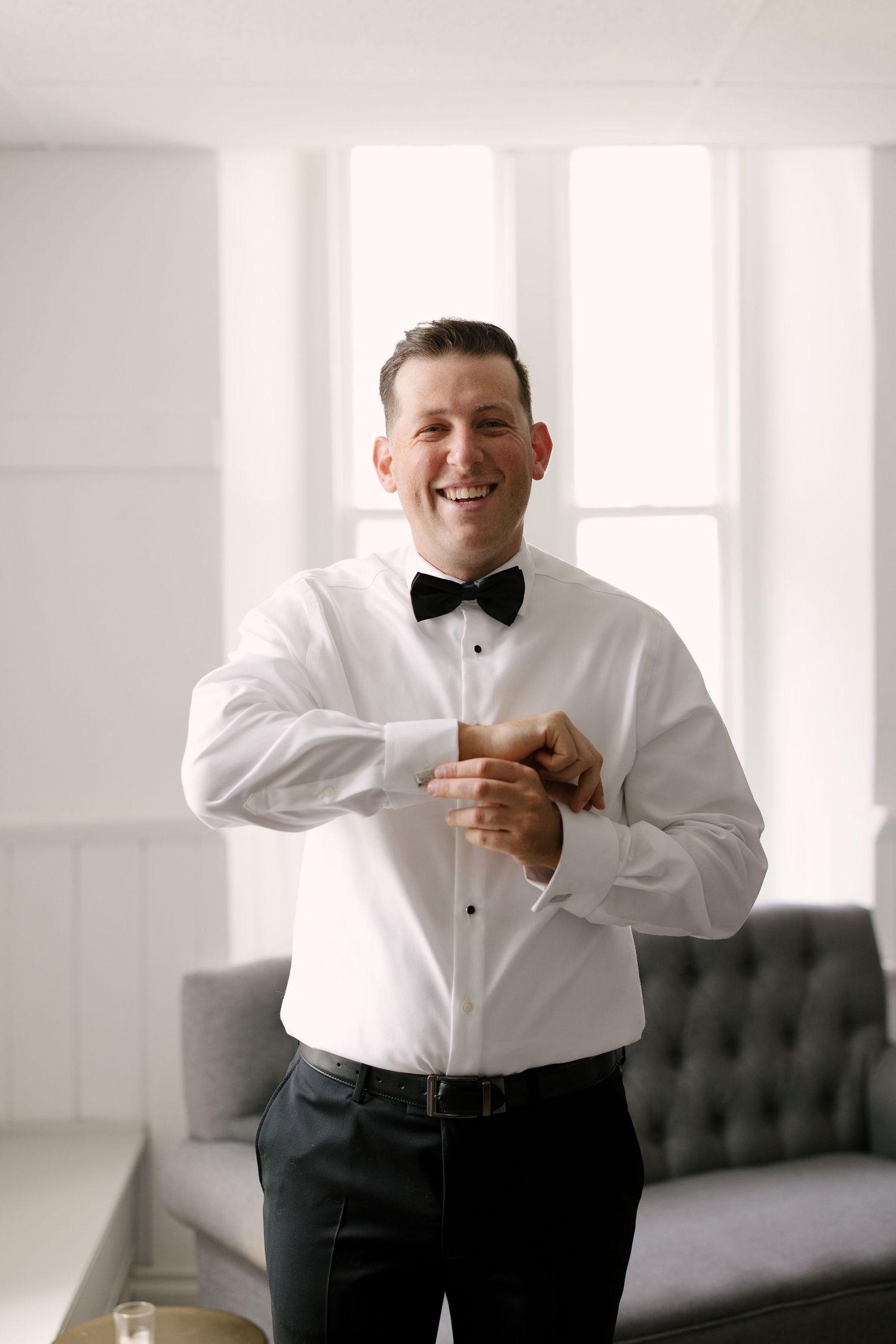 Smiling groom in white tuxedo shirt and black bow tie adjusts his cufflinks while getting ready for wedding.