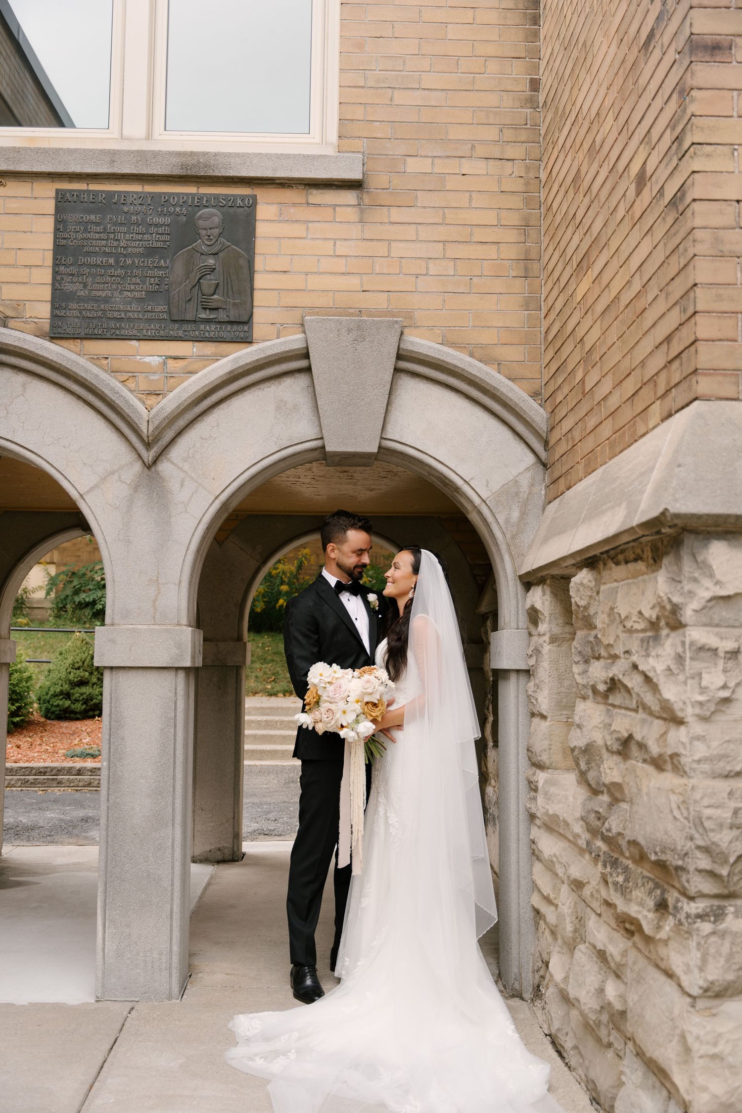 Elegant bride and groom posing under stone arches outside a classic brick church.