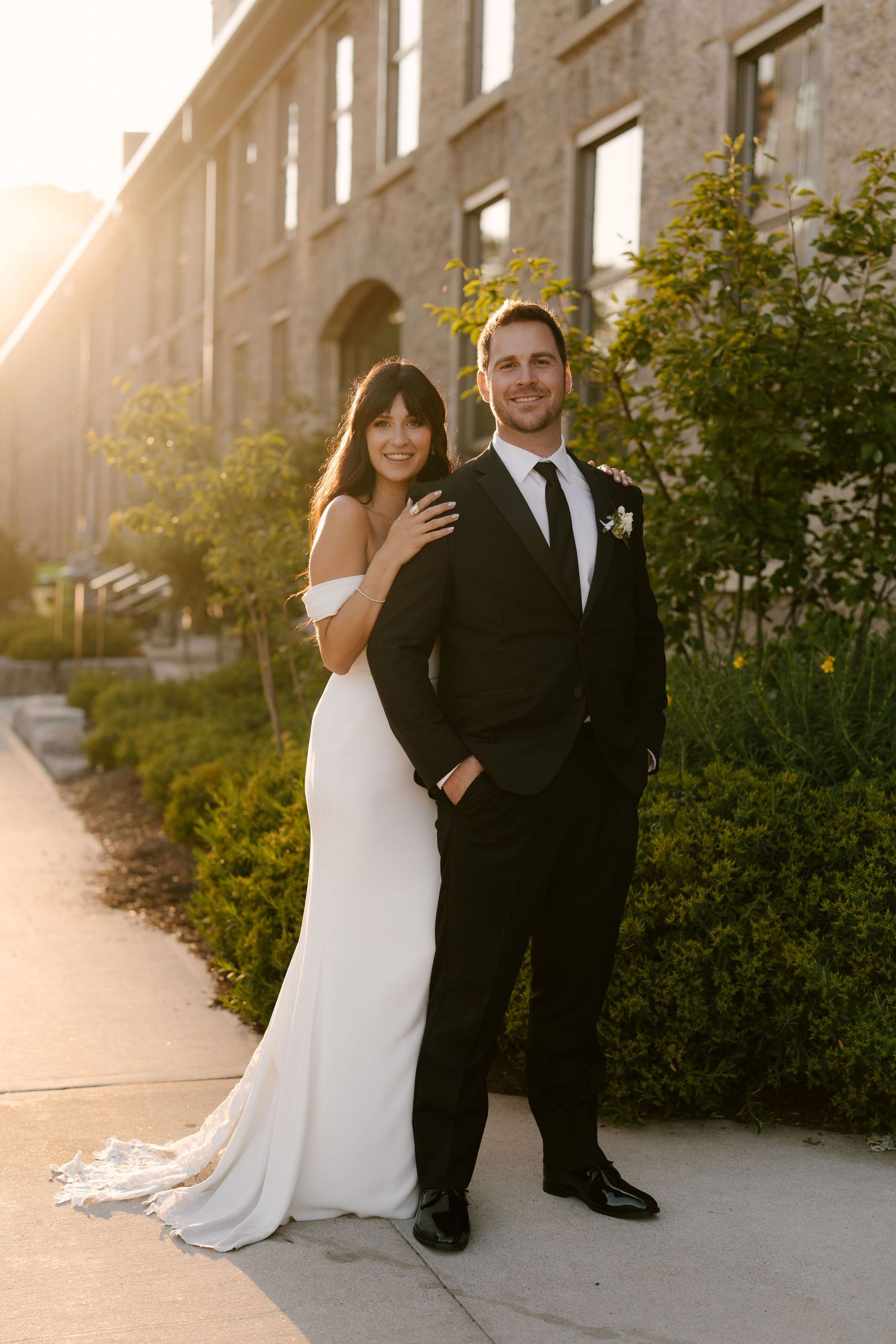 Bride and groom posing together at sunset near Tapestry Hall building, she in white gown, he in classic black tuxedo.