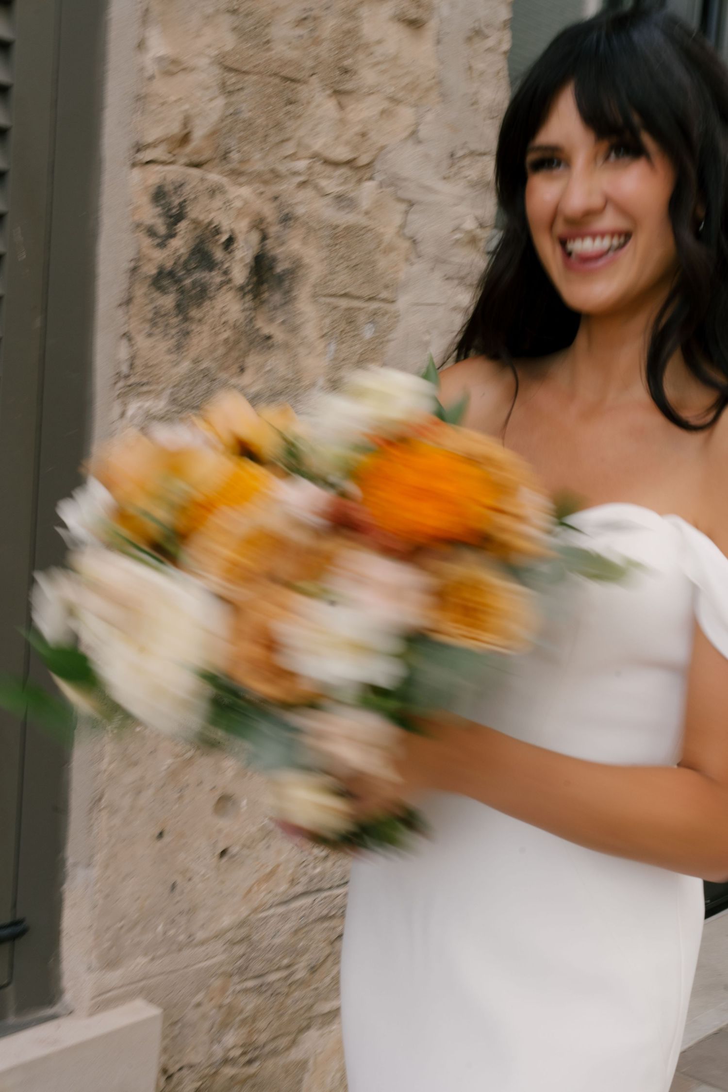 Joyful bride in white gown laughs while holding a blurred orange and white floral bouquet against a stone wall.