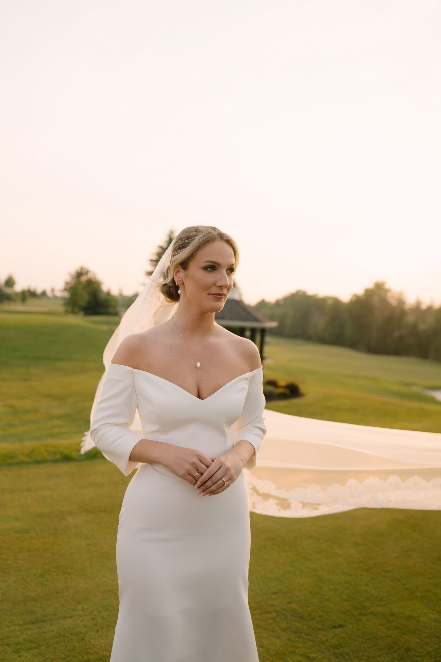 Elegant bride in off-shoulder white gown with lace veil flowing in breeze on green Whistle Bear golf course at golden hour.