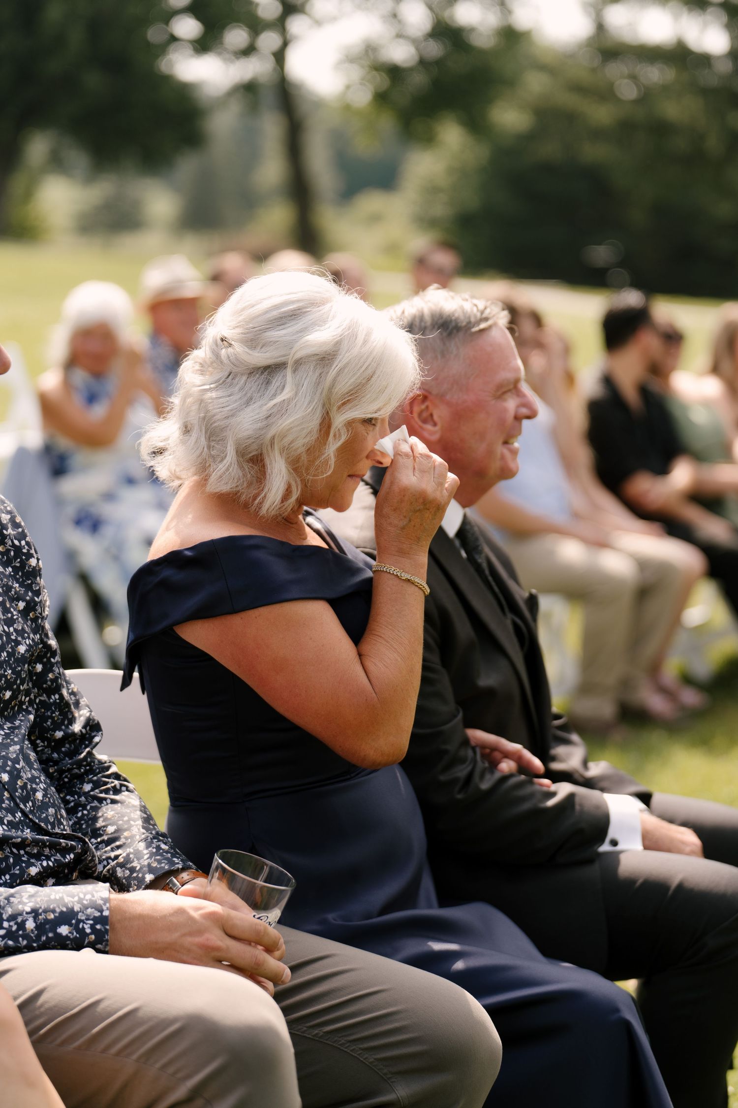 Emotional wedding guest in navy dress dabbing tears during an outdoor garden ceremony.