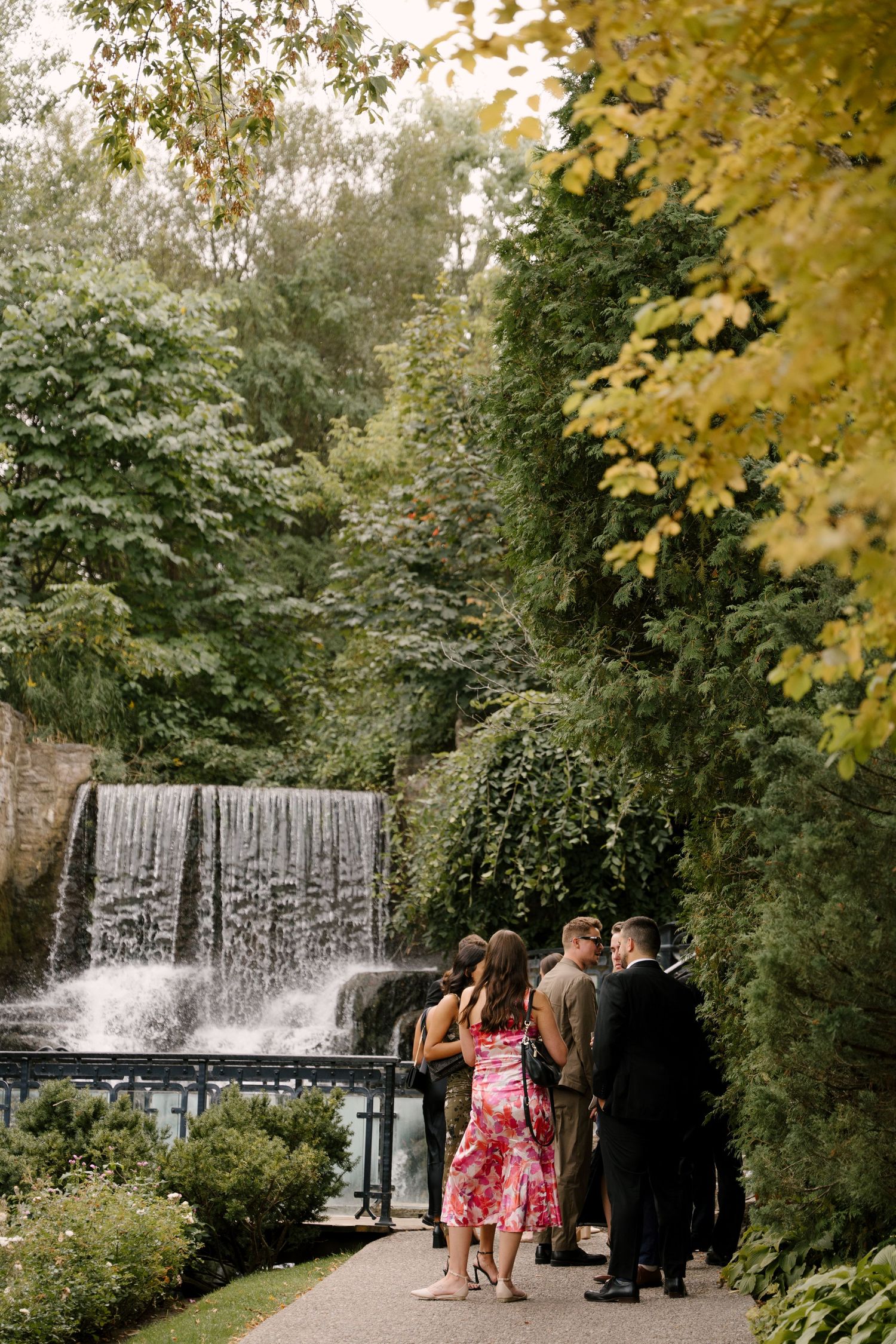 Wedding guests gathered near a scenic waterfall surrounded by lush greenery at an outdoor Ancaster Mill venue.