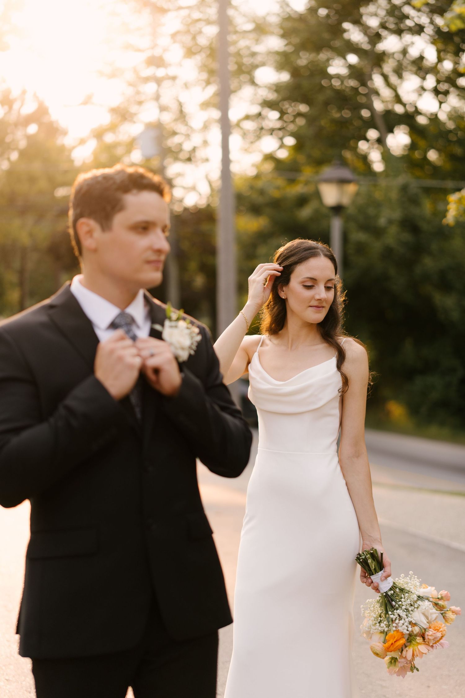 Bride in white gown holding bouquet and groom in black suit stand outdoors during golden hour sunset.