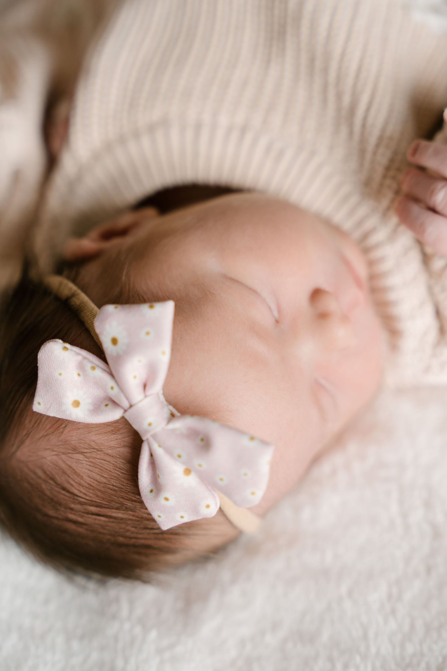 Sleeping newborn baby girl wearing pink polka dot bow headband on white fluffy blanket.