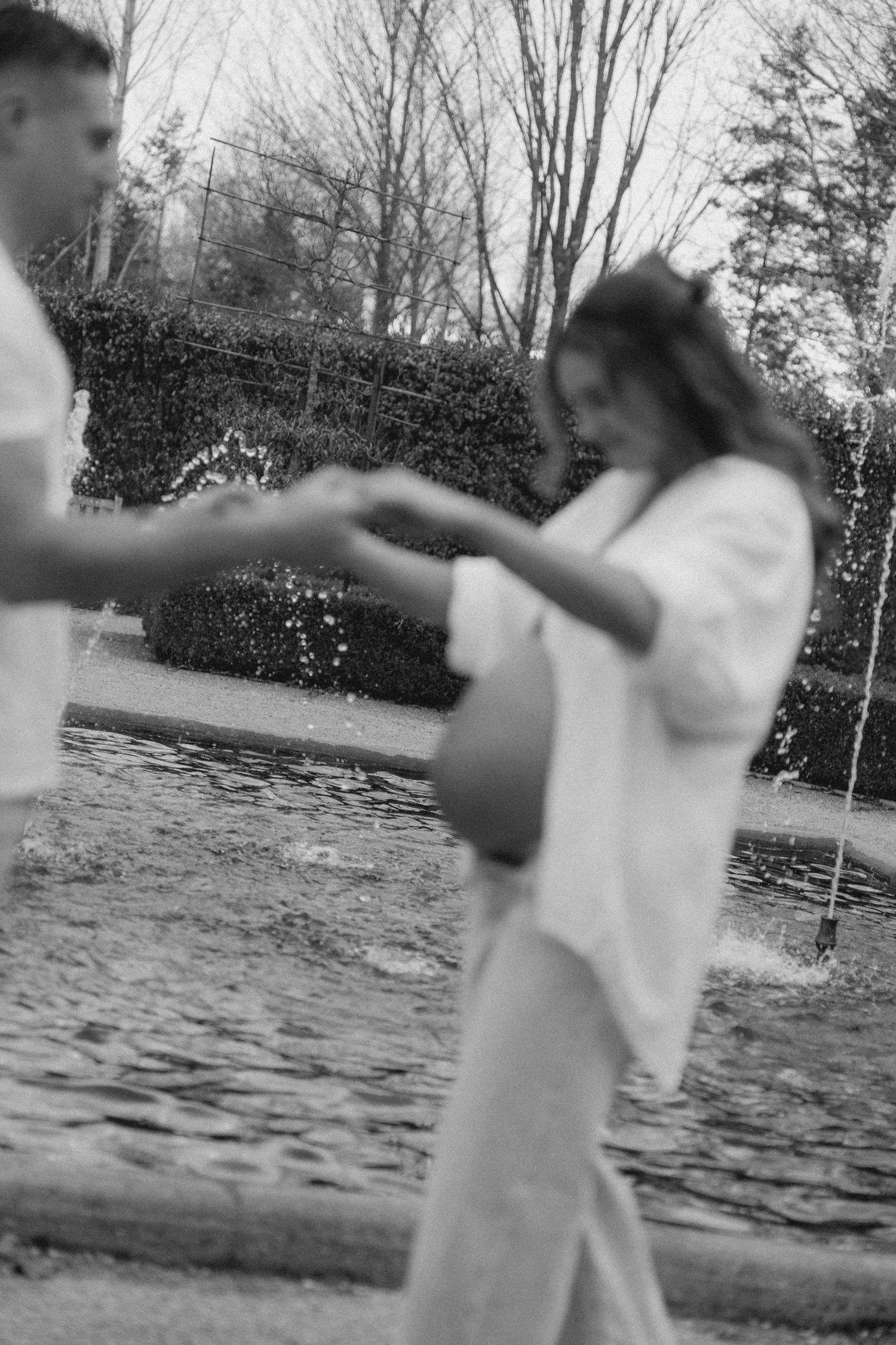 Black and white photo of a pregnant couple holding hands near a garden fountain.