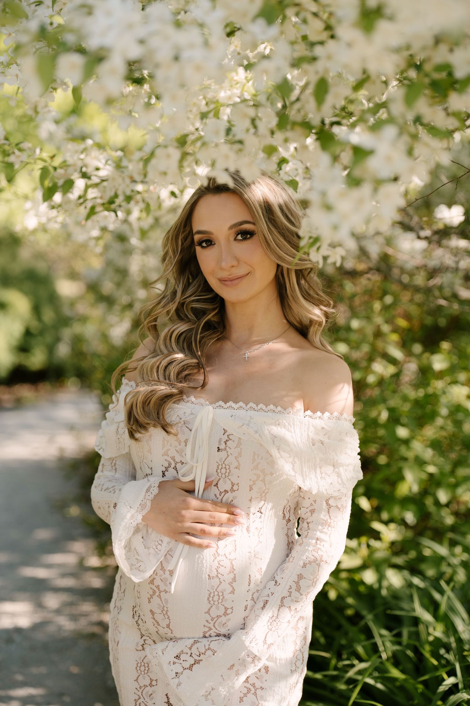 Pregnant woman in white off-shoulder lace dress posing near blooming white flowering trees.