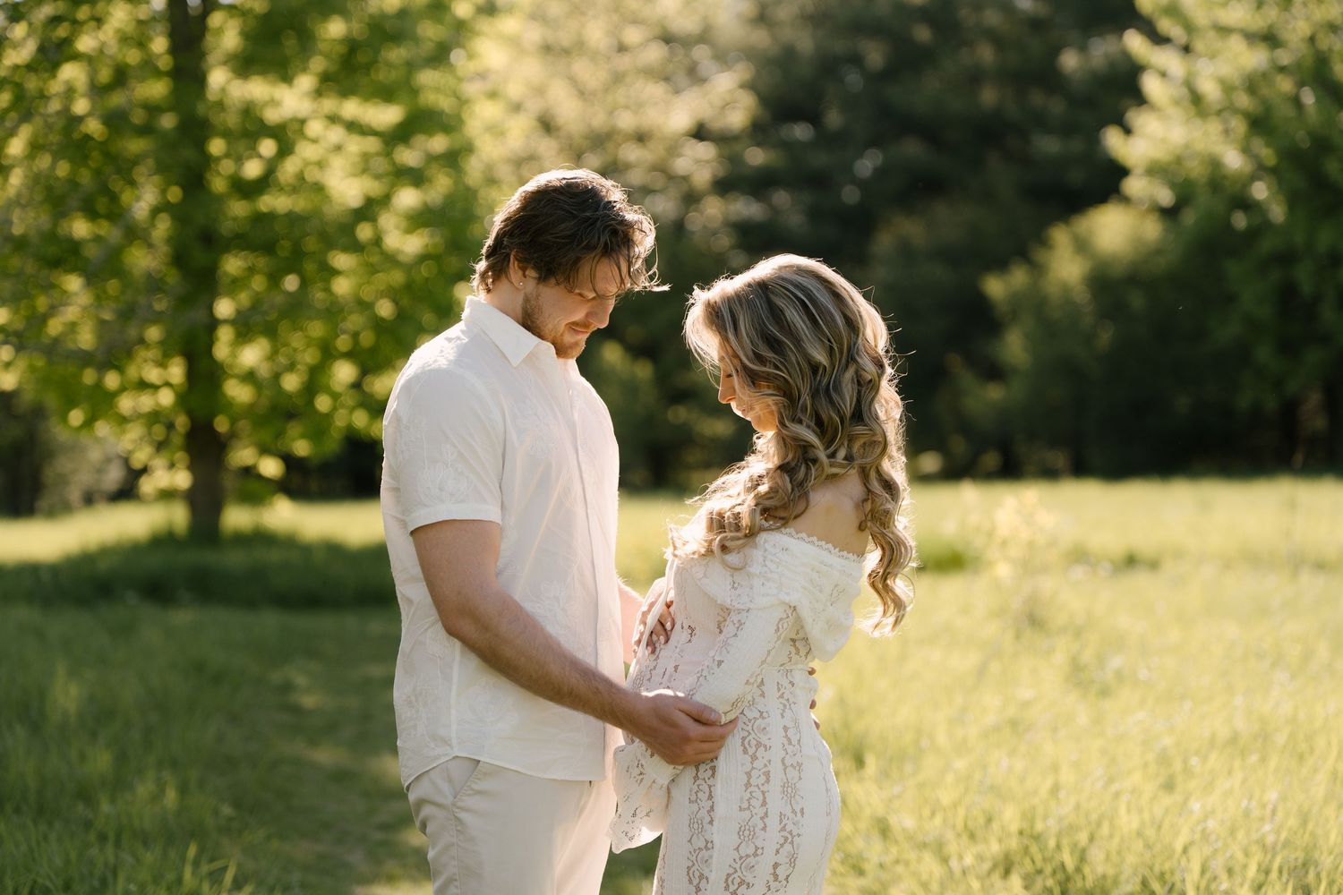 Pregnant couple holding hands and embracing in a sunlit green park, woman in white lace maternity gown.